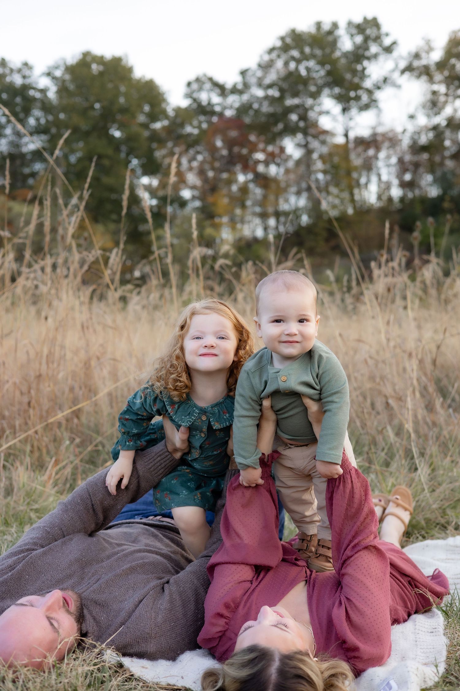 Two children lay together on a blanket in a field of tall grass during golden hour.