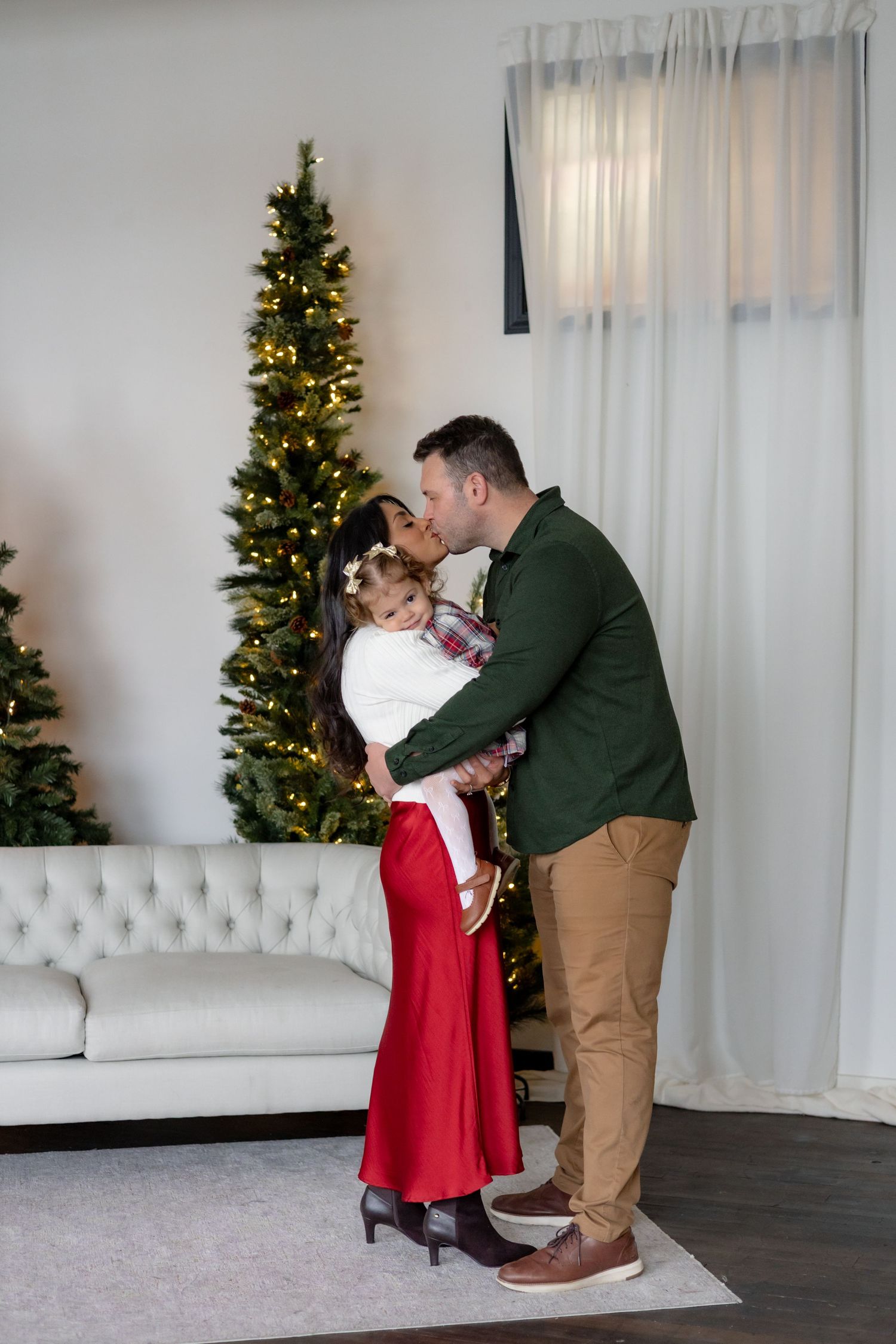 A family shares an intimate moment by a lit Christmas tree in their living room with white decor.
