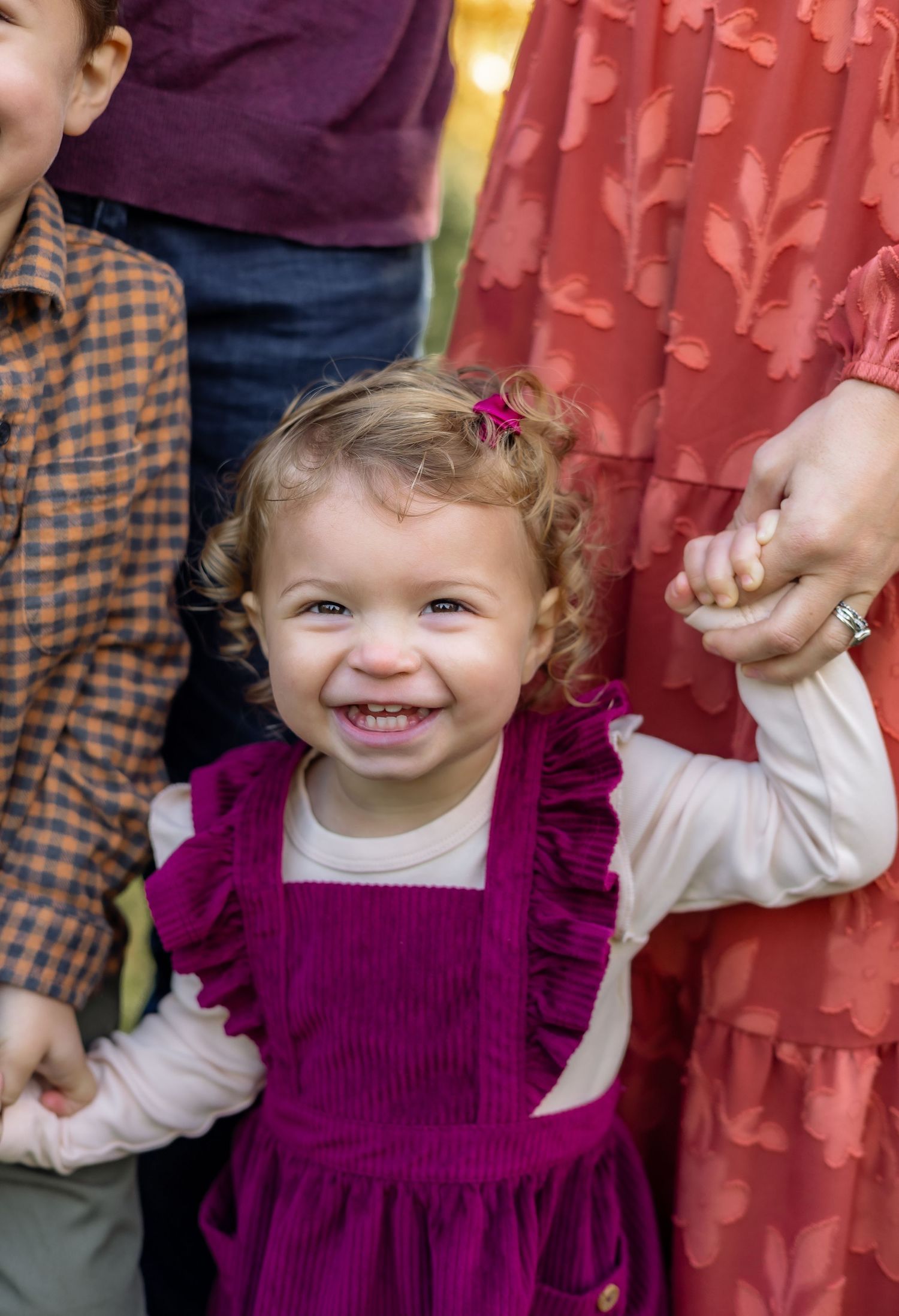 A child in a purple dress smiles and plays while standing against a red curtain background.