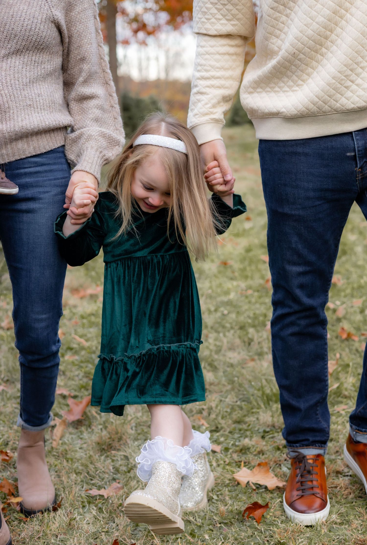Small child in emerald green dress swings playfully between two adults holding hands on autumn grass.