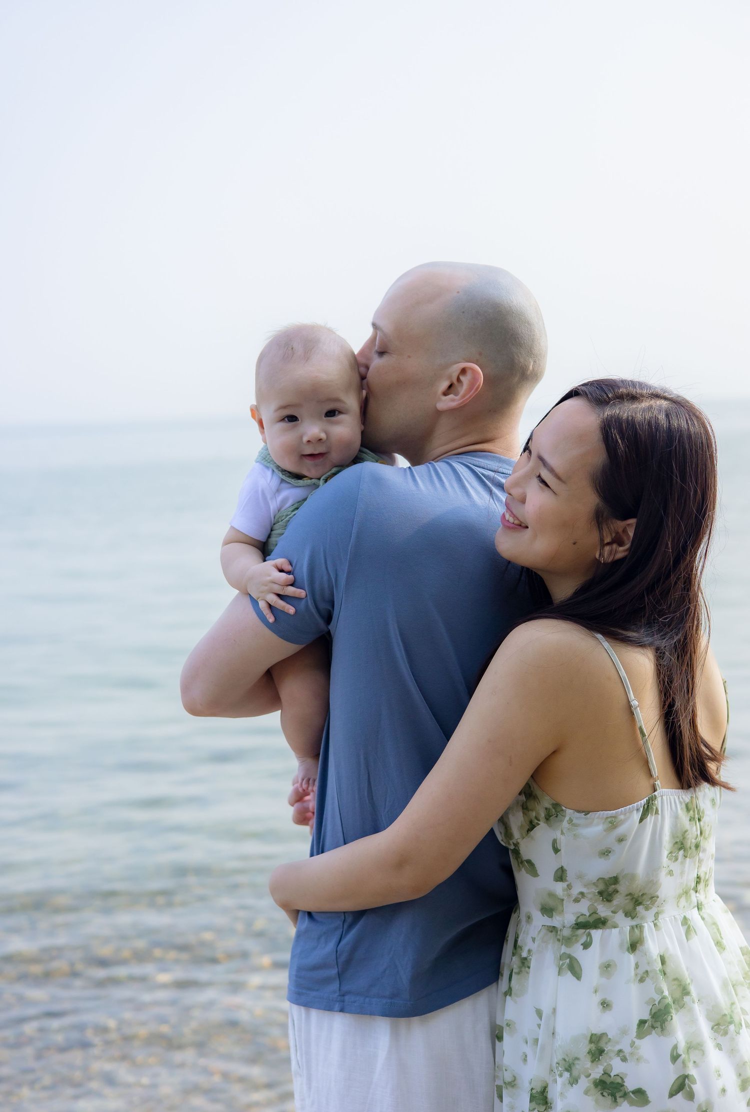 A couple shares tender moments with their baby while standing near a serene waterfront on a bright day.