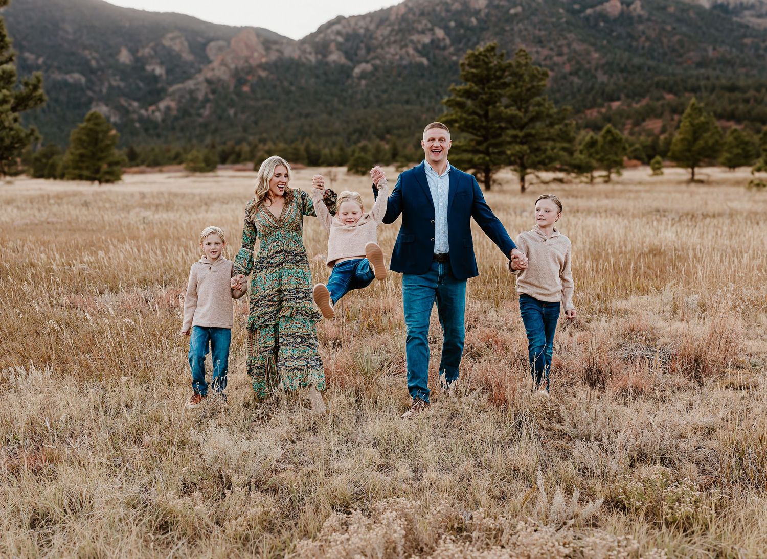 A group of four people hold hands while walking through a golden autumn field with mountains in the background.