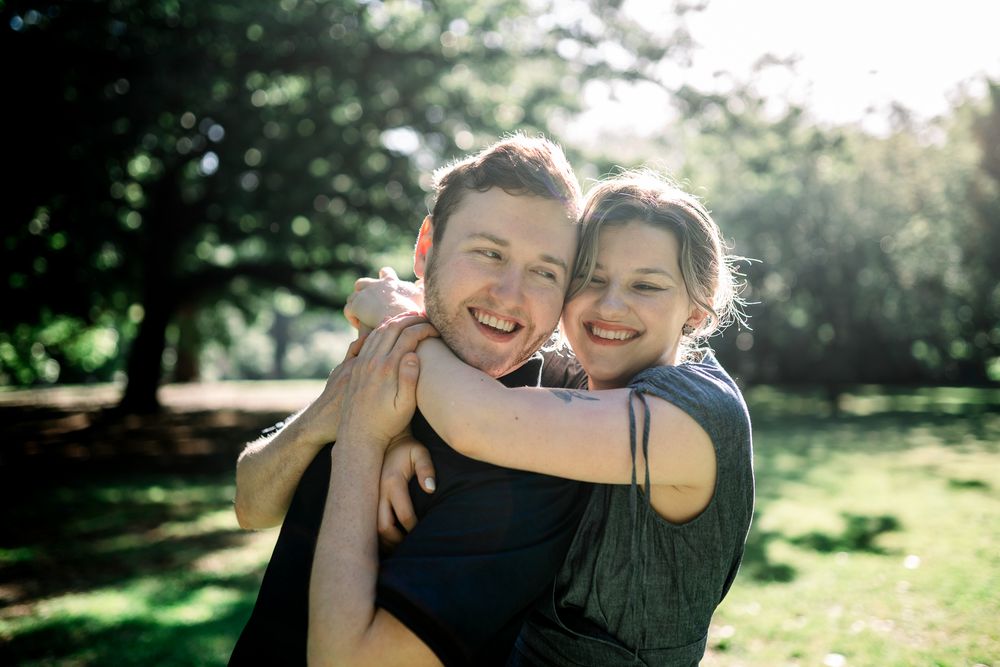 Two people share a warm embrace and laugh together in a sunny park with green trees taken by Melbourne couples photographer
