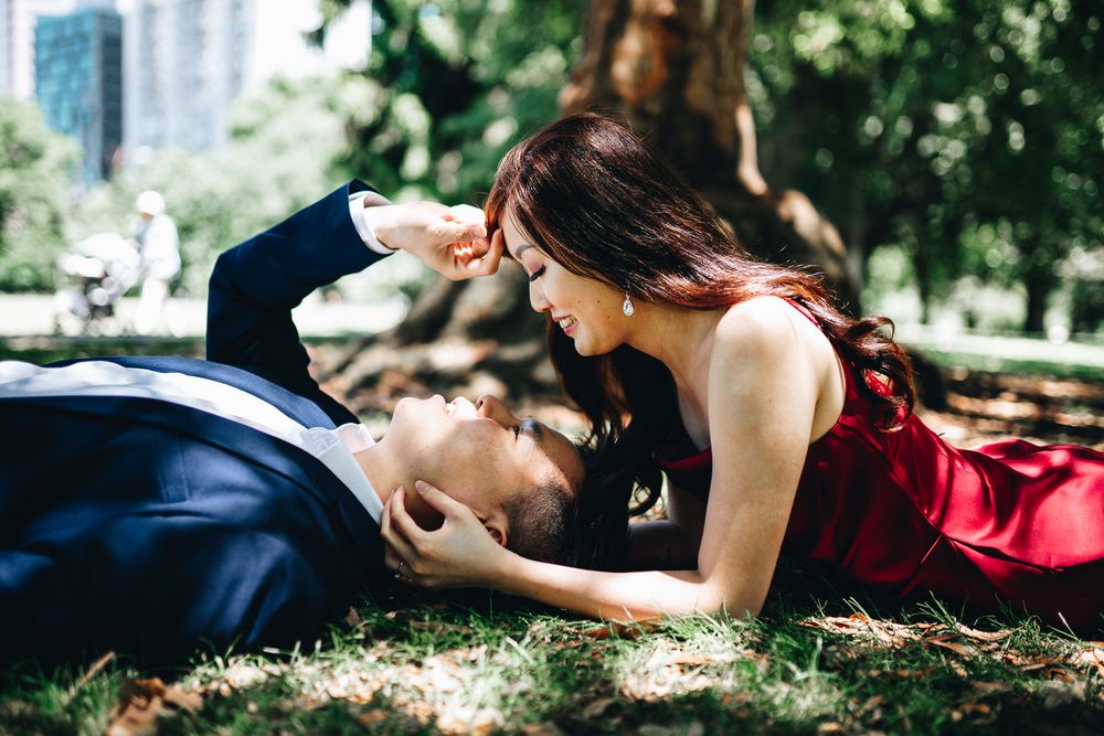 A romantic couple in formal attire lying on grass in a park with cityscape visible in background taken by Melbourne couples photographer