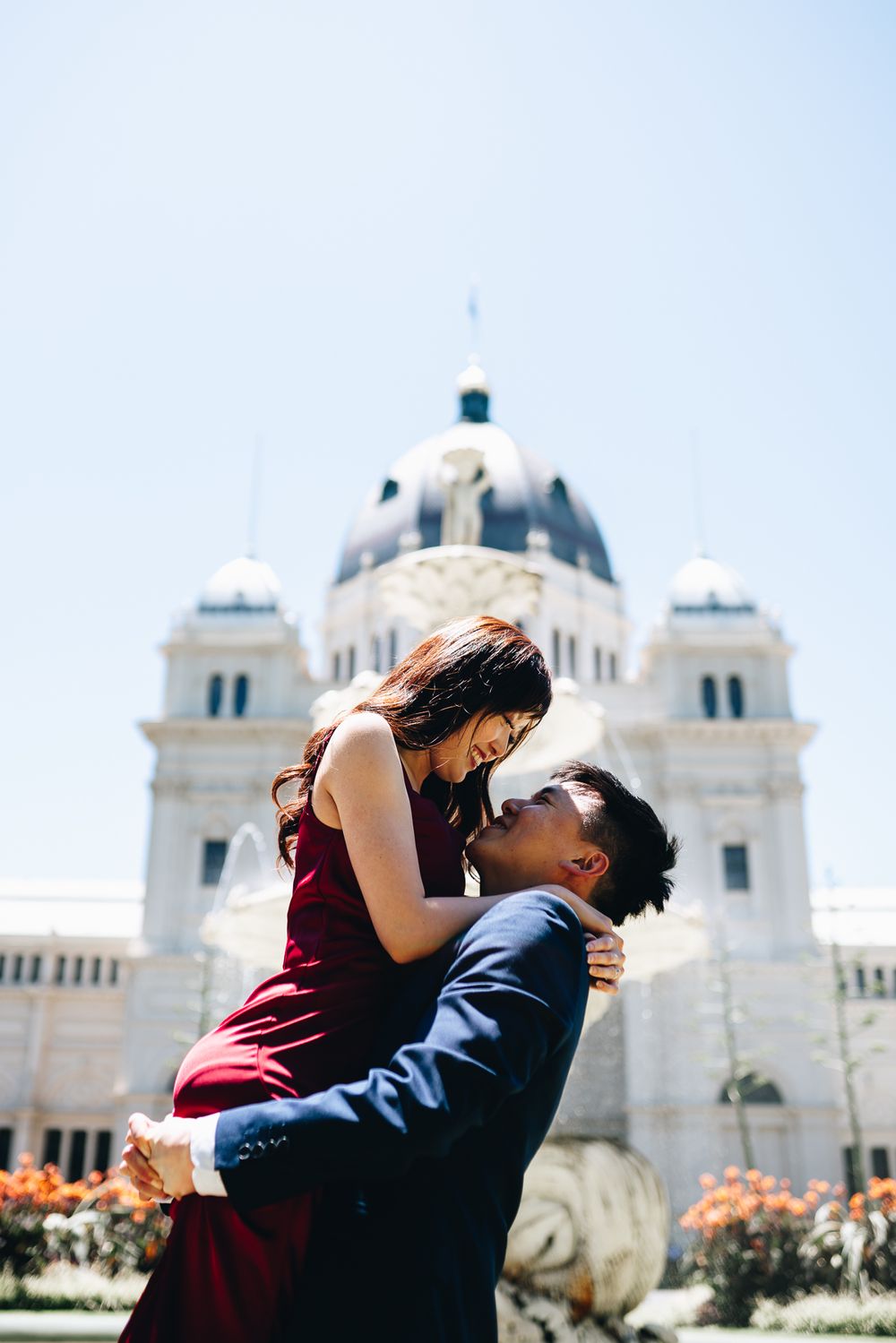 A couple embraces in front of an ornate domed building with white architecture and blue sky taken by Melbourne couples photographer