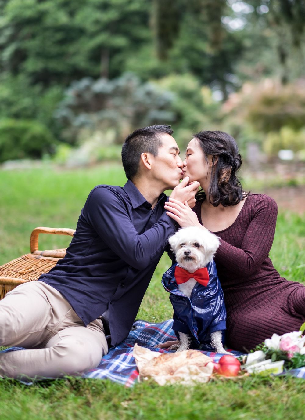 A couple shares a kiss during a picnic in a park with their small white dog wearing a bow tie taken by Melbourne couples photographer.
