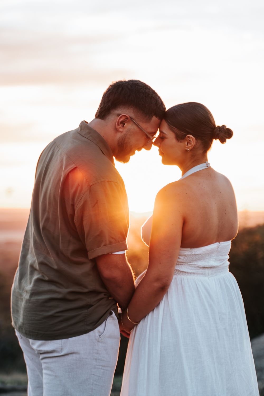 Couple holding hands and touching foreheads at golden sunset, man in gray shirt and woman in white halter dress.