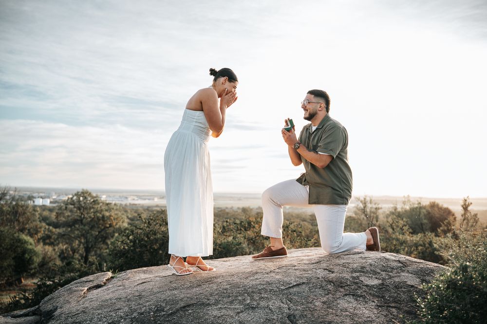 Man proposing on one knee to woman in white dress on rocky hilltop at sunset with scenic landscape behind them.