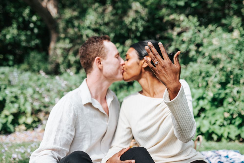 A newly engaged couple showing off her engagement ring while kissing in the background during a surprise proposal in Melbourne.