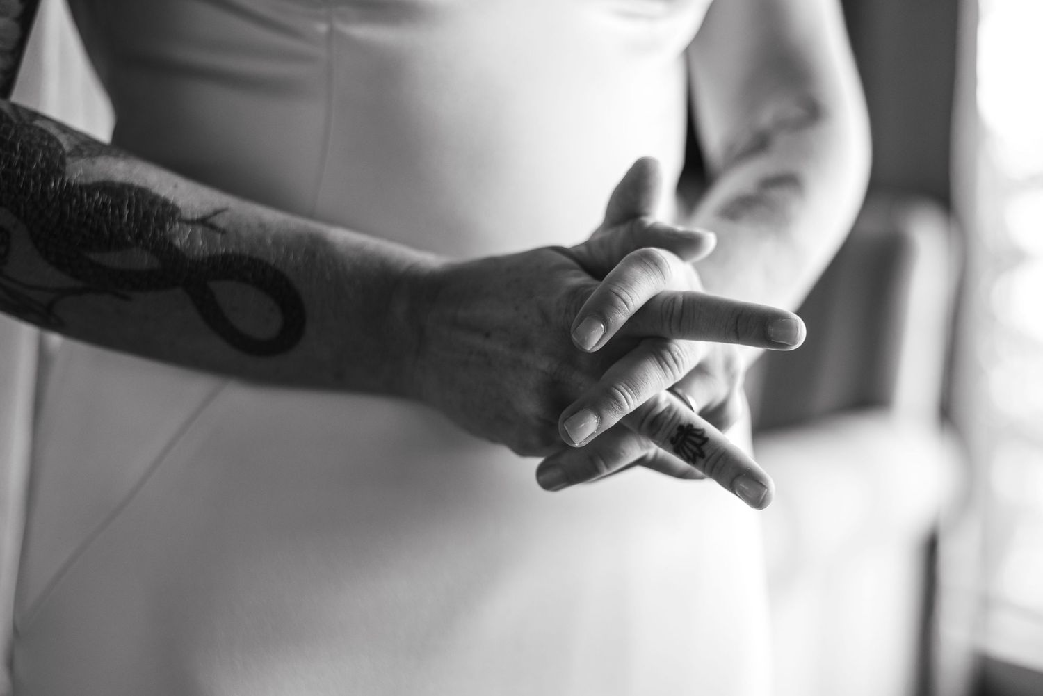 Black and white close-up photo of tattooed hand wearing rings in elegant dress.