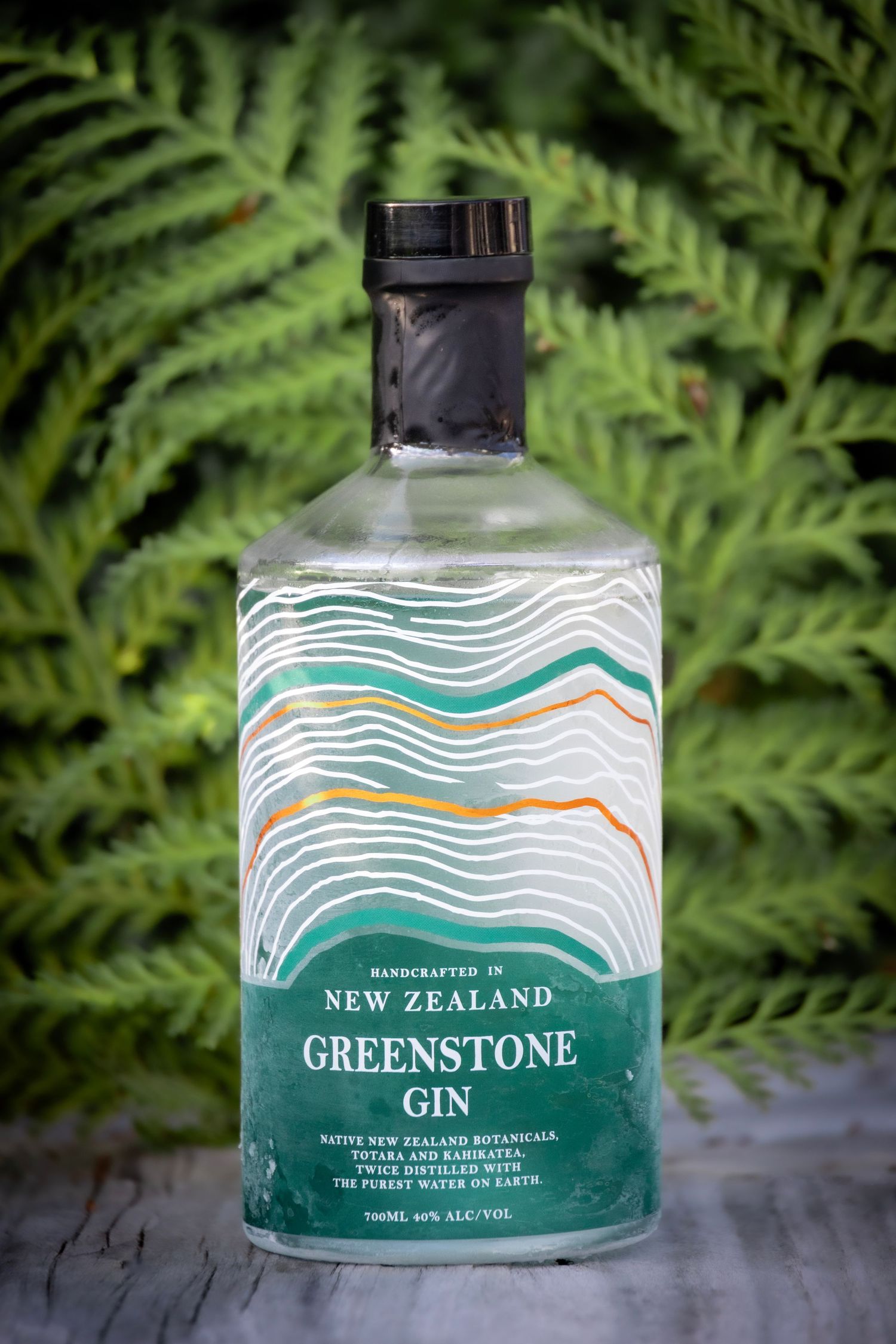 Bottle of New Zealand Greenstone Gin with a wavy label design, set in front of lush green ferns on a natural background.