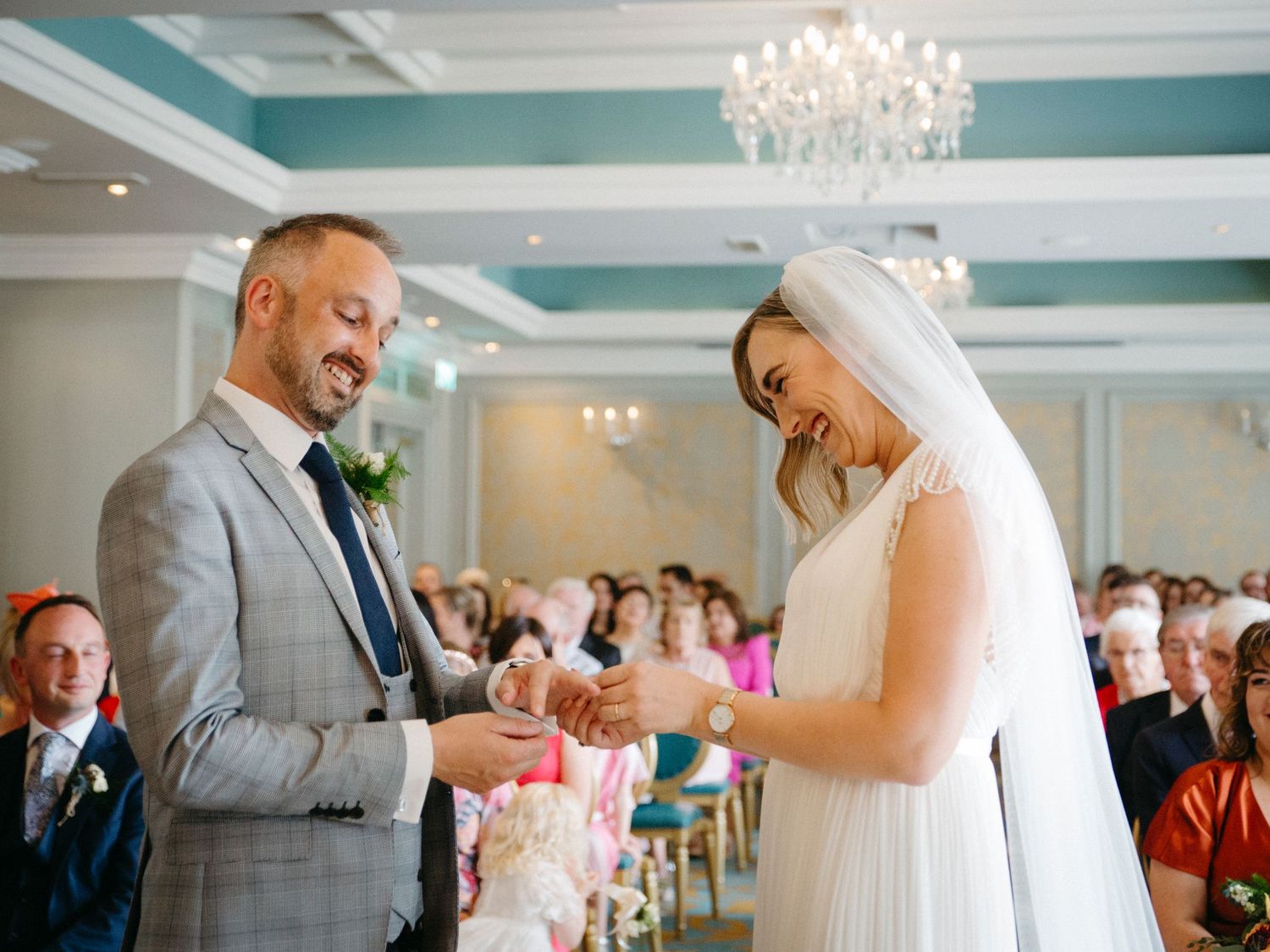Guests attend an indoor wedding ceremony seated in elegant vintage chairs on patterned carpet under warm lighting.
