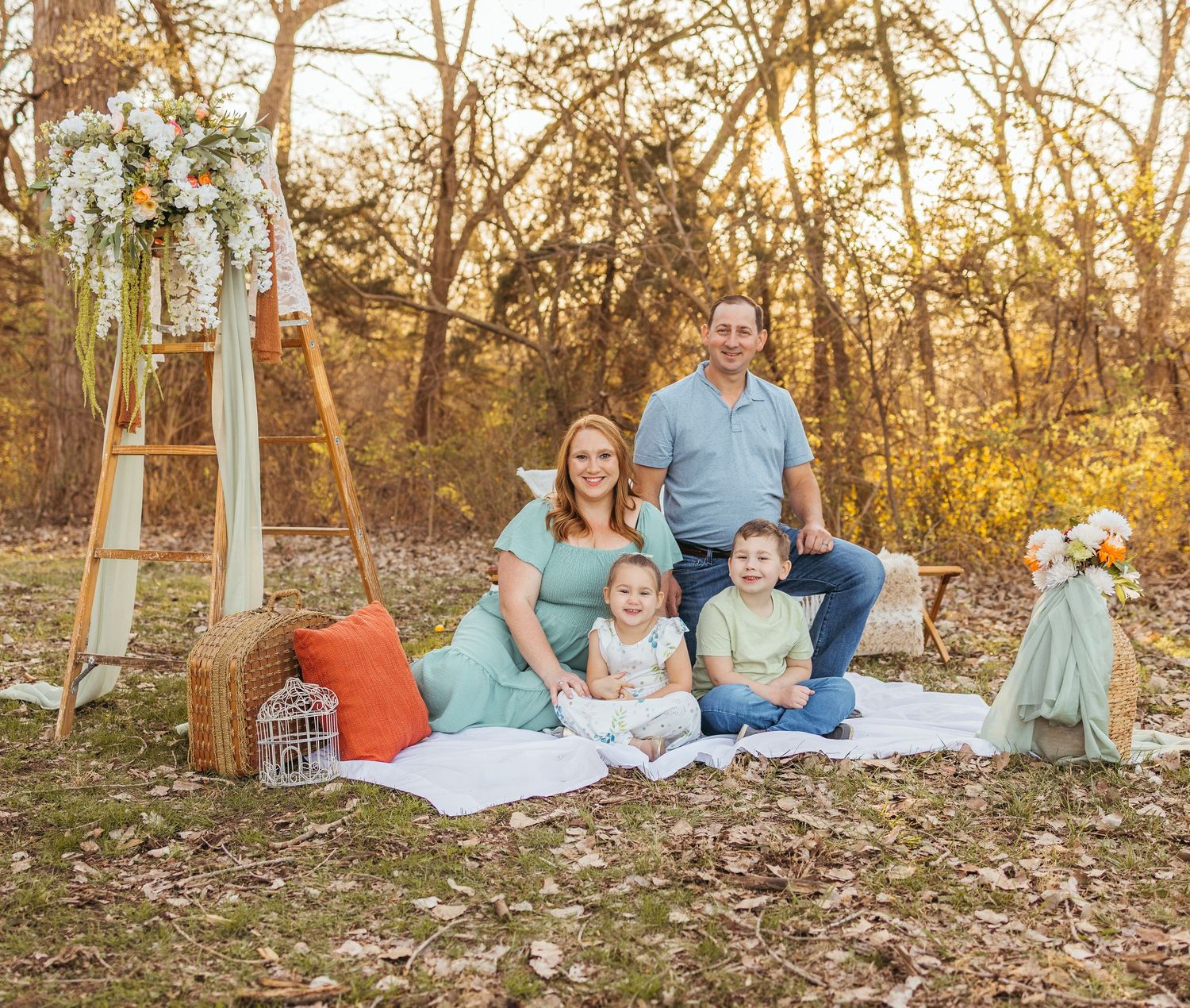 Family sitting on a picnic blanket in a woodsy autumn setting with vintage decor including a ladder and throw pillows.
