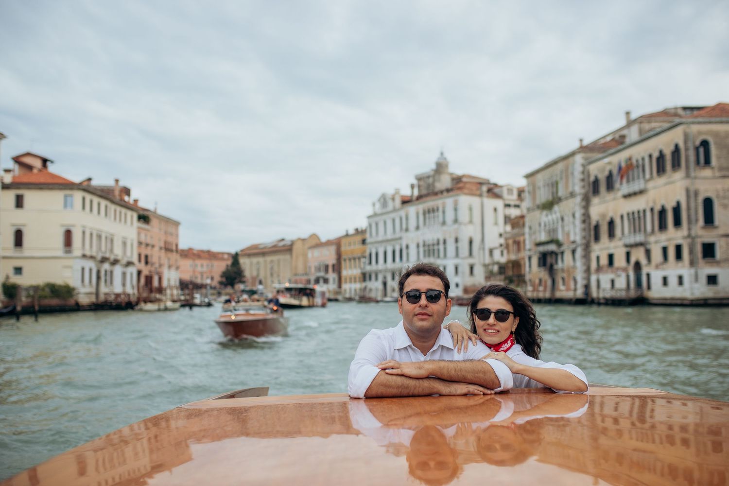 Two tourists enjoy views of historic Venetian buildings and canals while riding in a traditional wooden water taxi in Venice, Italy.