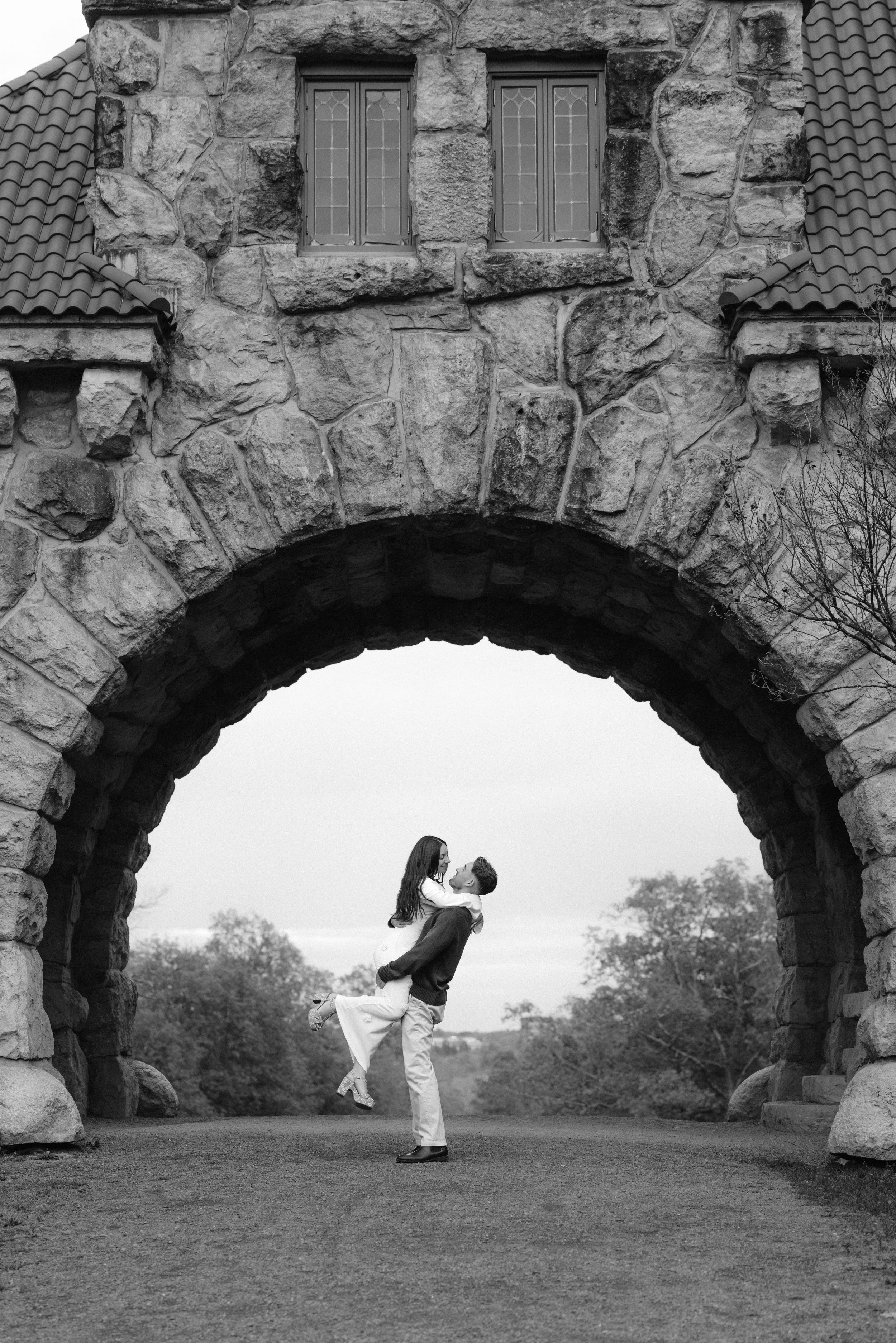 A romantic silhouette of a couple embracing under a rustic stone archway in black and white photography.