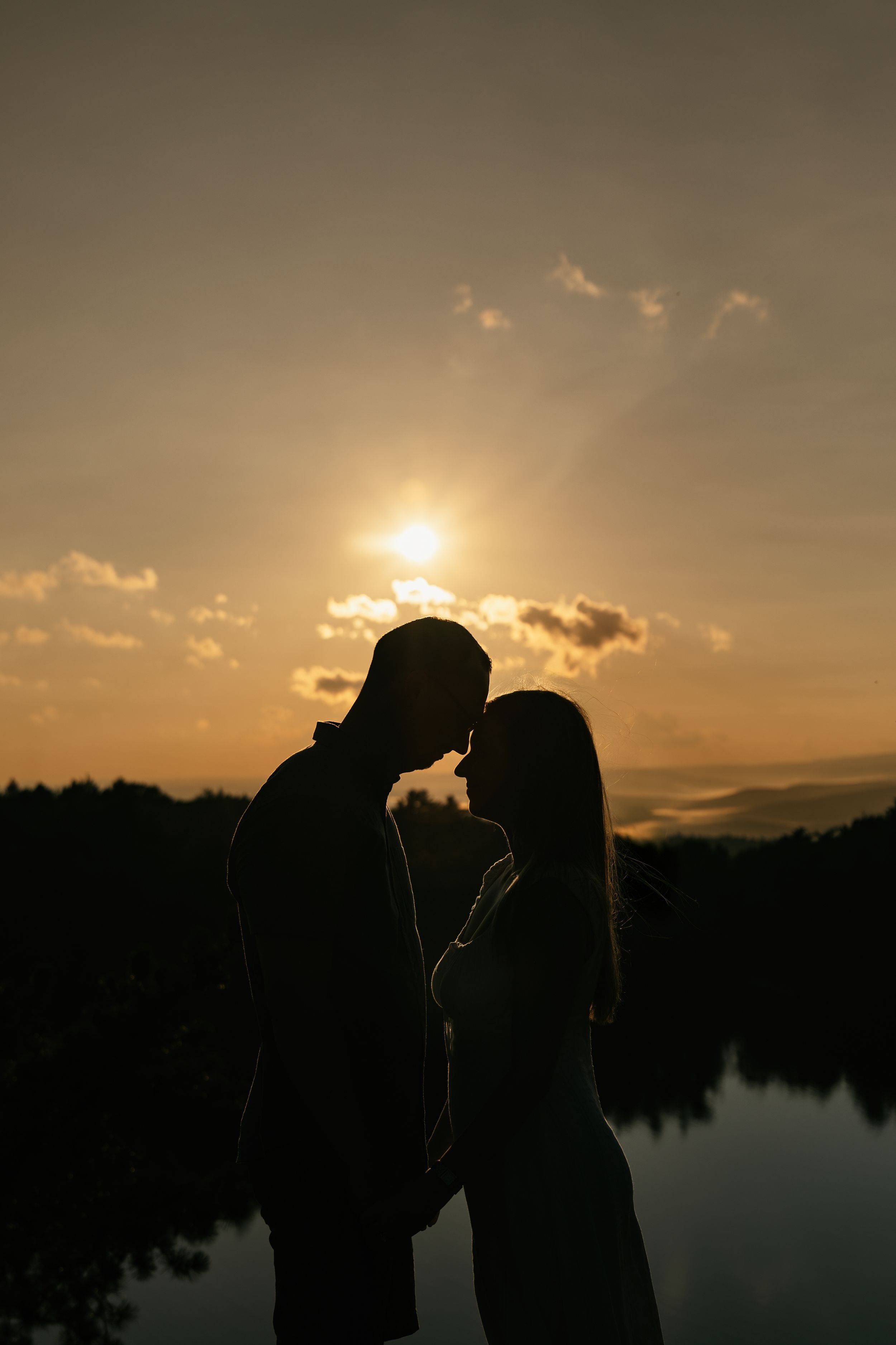 Silhouette of a romantic couple standing close together against a golden sunset over a tranquil lake.