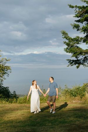 A couple walks hand in hand through a grassy hillside with dramatic cloudy skies and evergreen trees in the background.