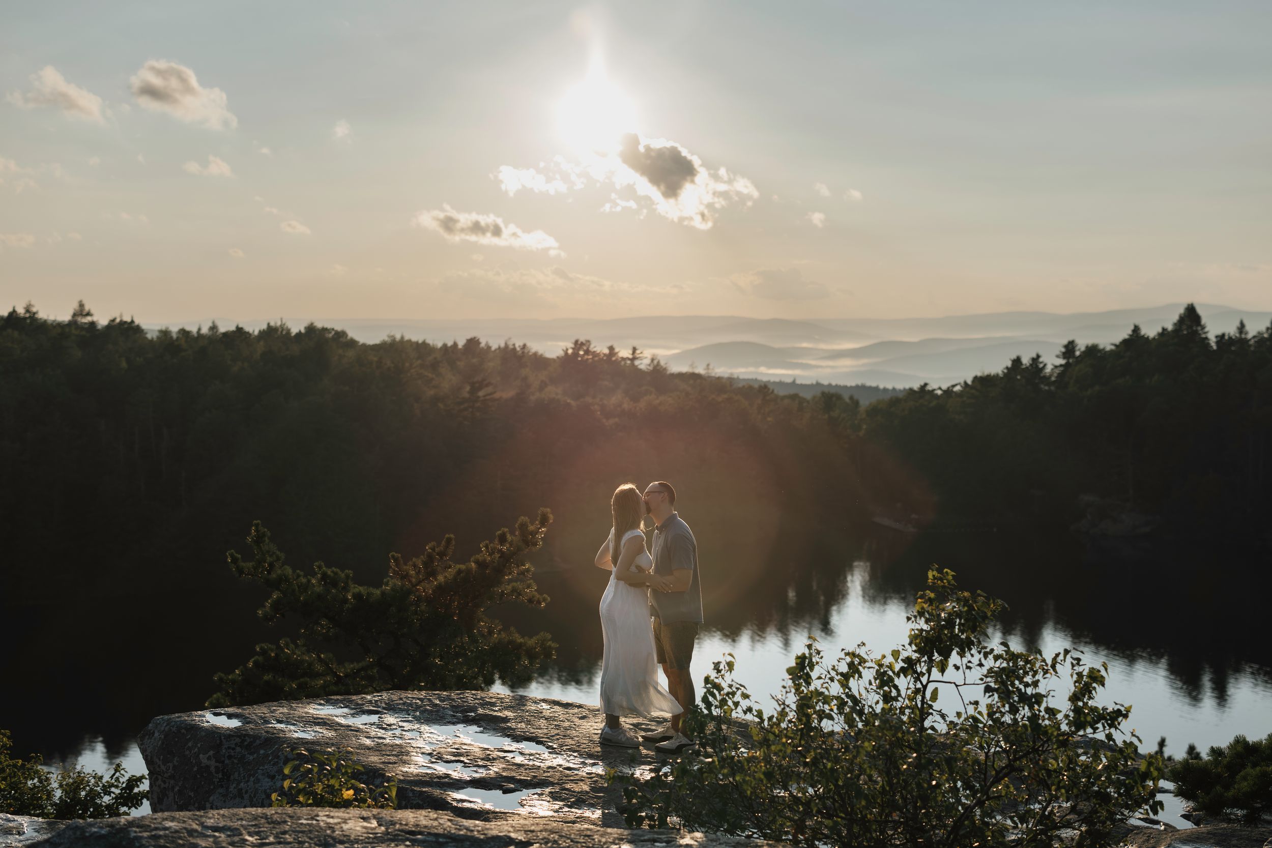 A couple stands on a rocky cliff overlooking a serene lake at sunset with mountains in the background.