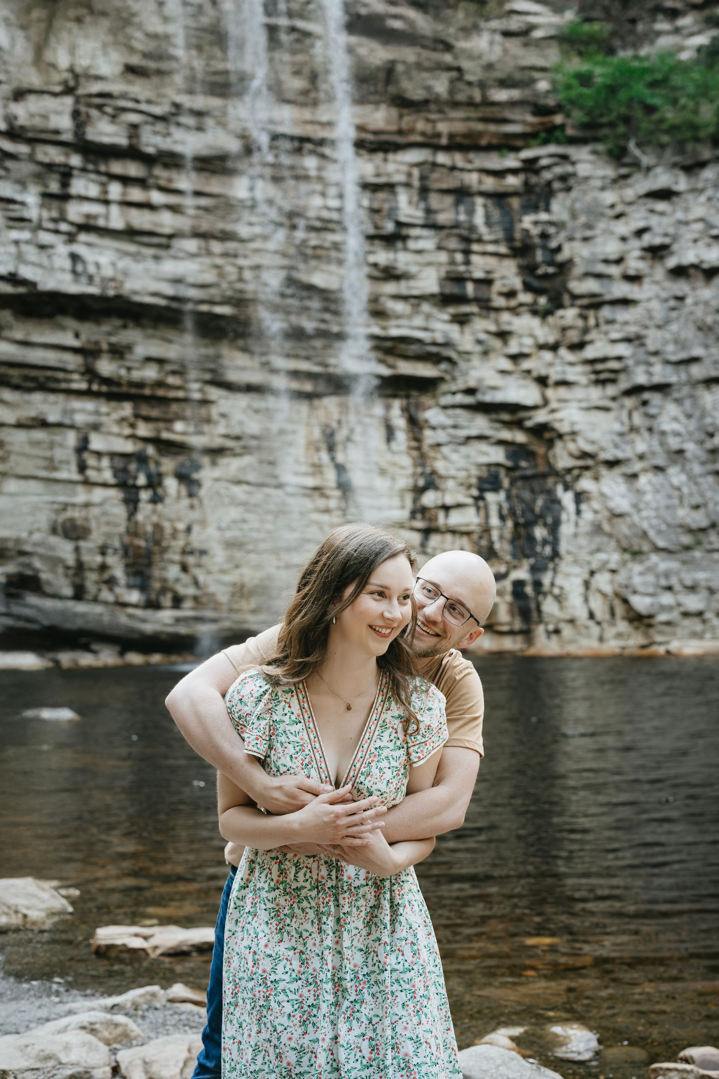 A couple embraces by a scenic waterfall and rocky cliff face at a natural swimming hole.