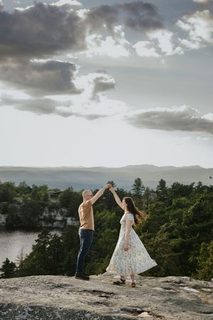 Two people dance together on a rocky cliff overlooking a scenic lake and forest landscape at sunset.