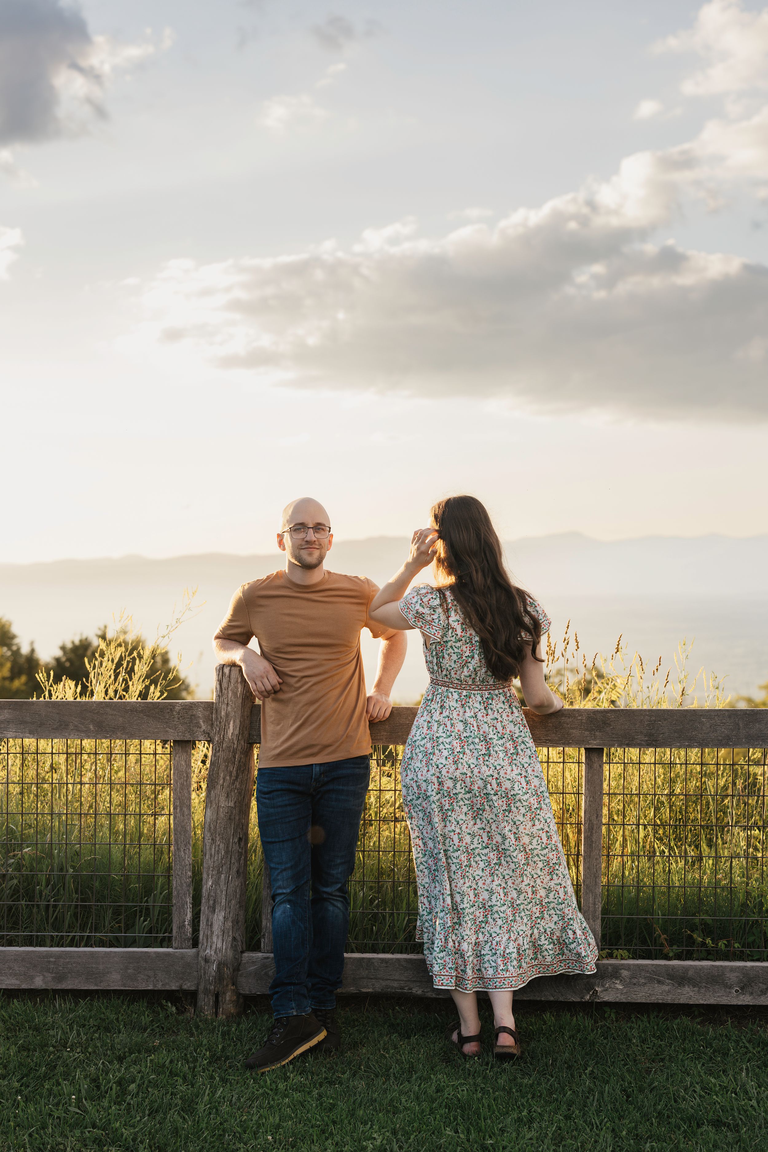 A couple sits together on a rustic wooden fence at sunset overlooking a pastoral countryside view.