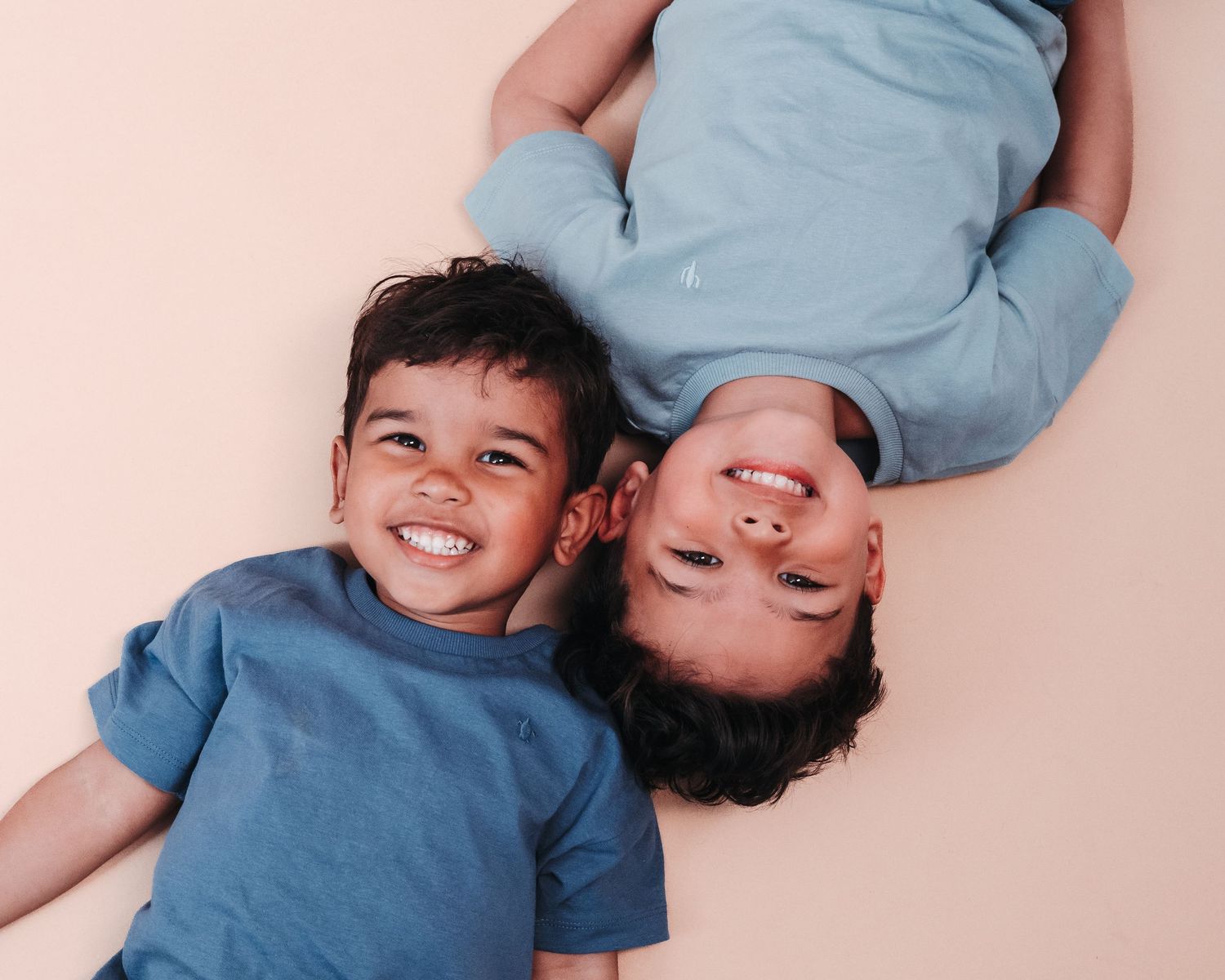 Two children in matching blue shirts lying head to head on pink background.