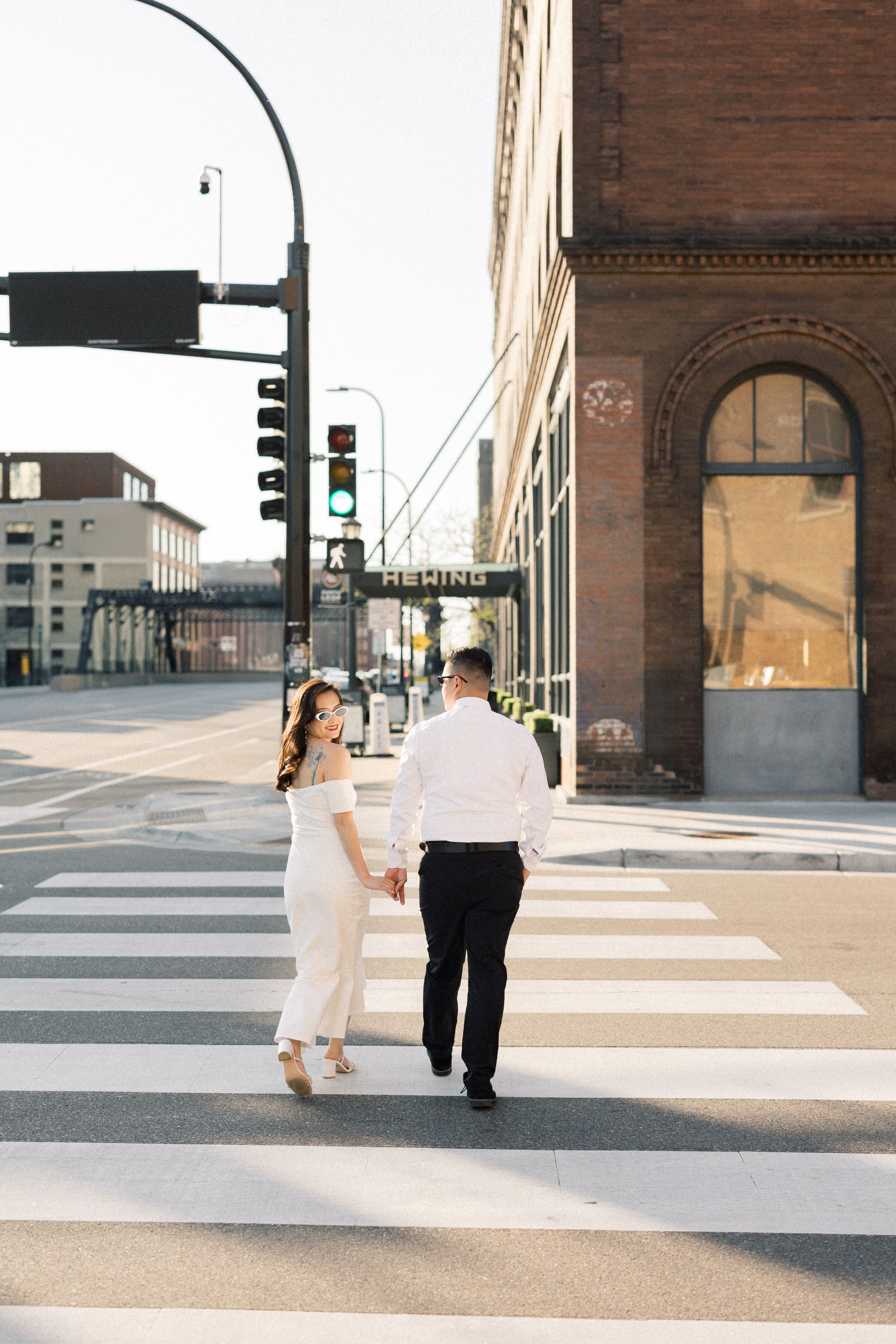 A couple holds hands while crossing a crosswalk in downtown at sunset with historic brick buildings in the background.