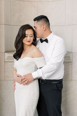 A romantic couple poses intimately against a stone wall, with one person in a white dress and another in formal attire.