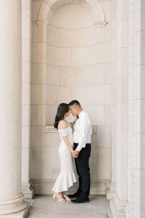 A couple shares an intimate moment in formal attire while standing between elegant stone columns in an alcove.