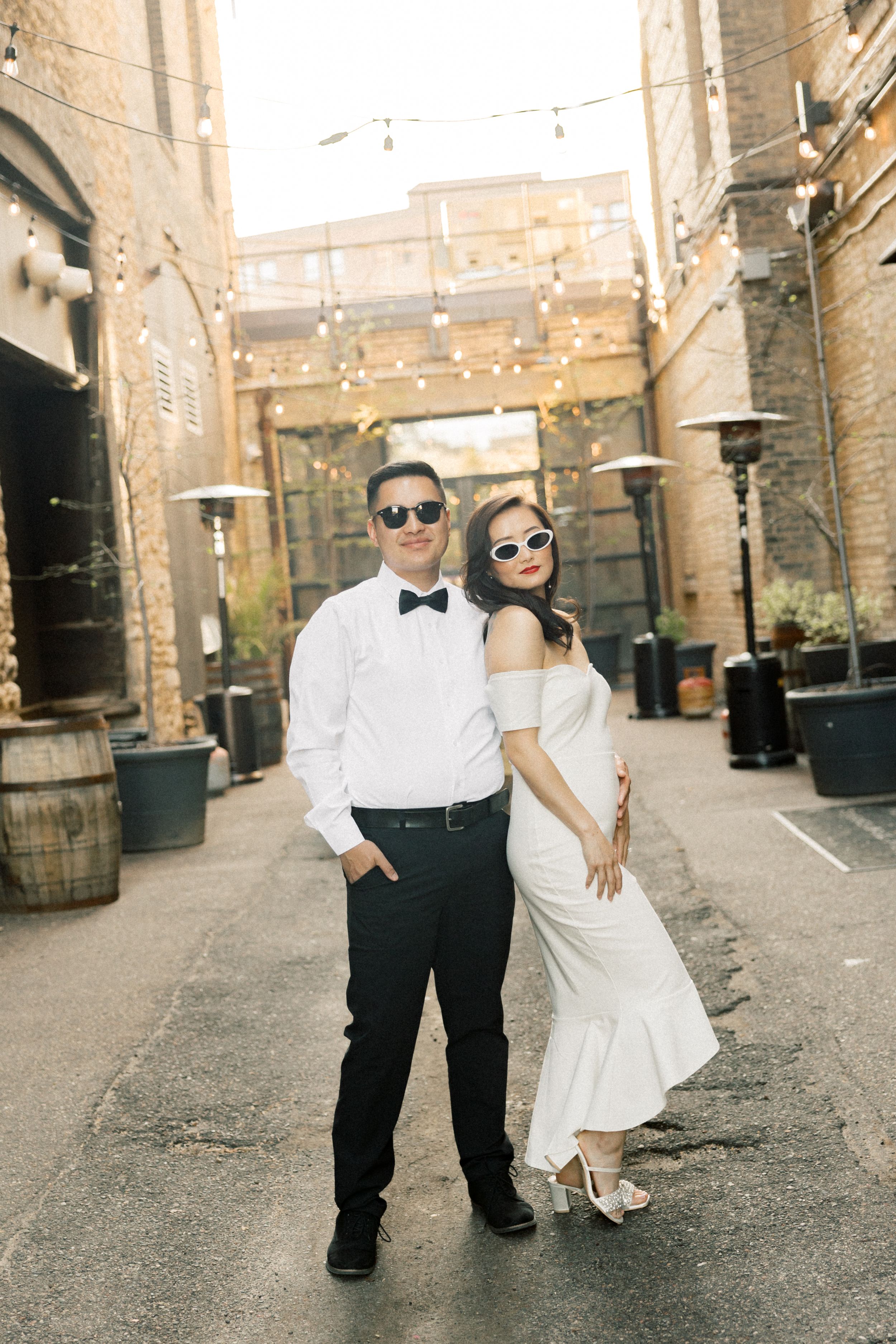 A couple in formal attire poses in an urban alley with string lights overhead and stone walls on either side.