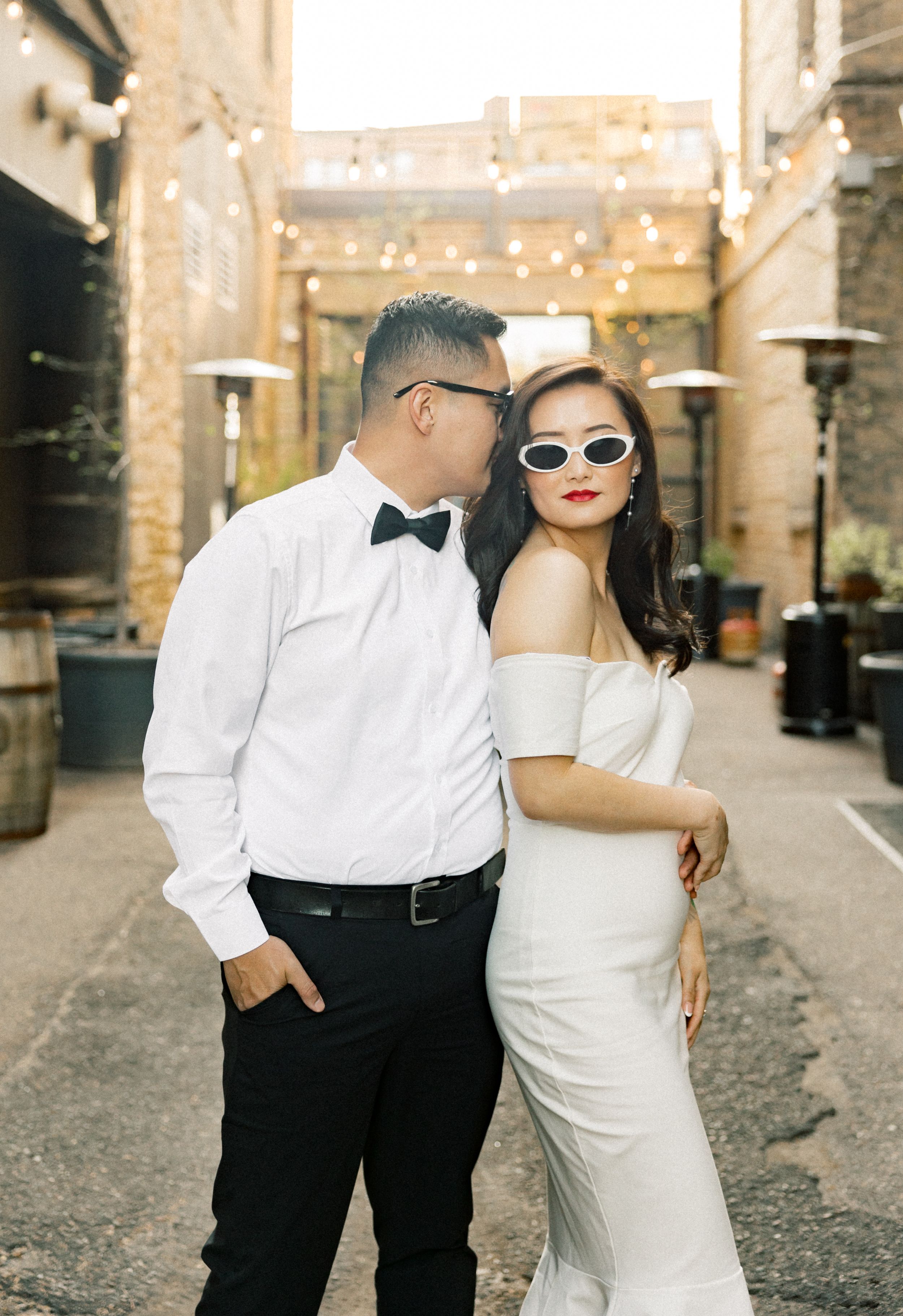 A couple poses for a wedding photo in an urban alley decorated with string lights, wearing formal attire and sunglasses.