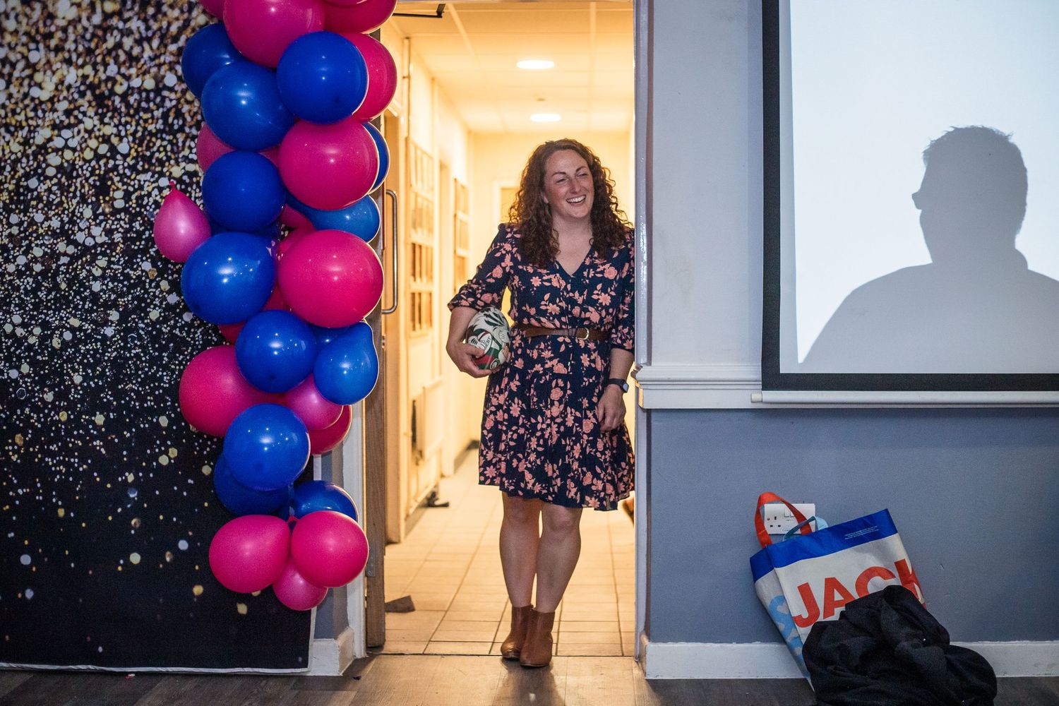 Woman wearing a dress, holding a rugby ball stands in a well lit doorway beside a balloon arch.