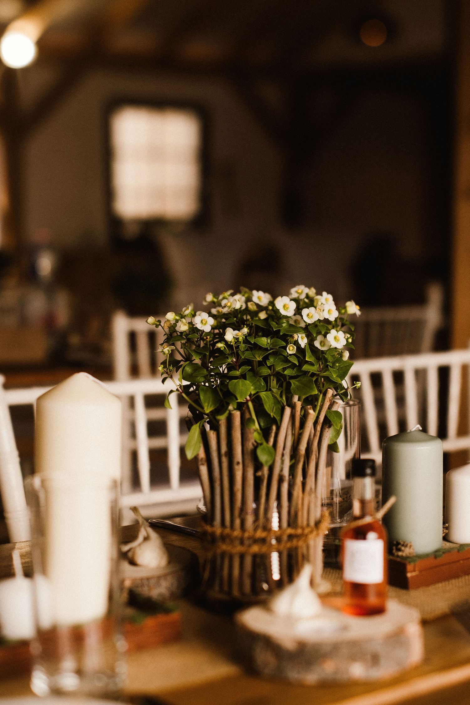 Rustic centerpiece with white flowers in a twig-wrapped vase surrounded by candles on an elegant table setting.