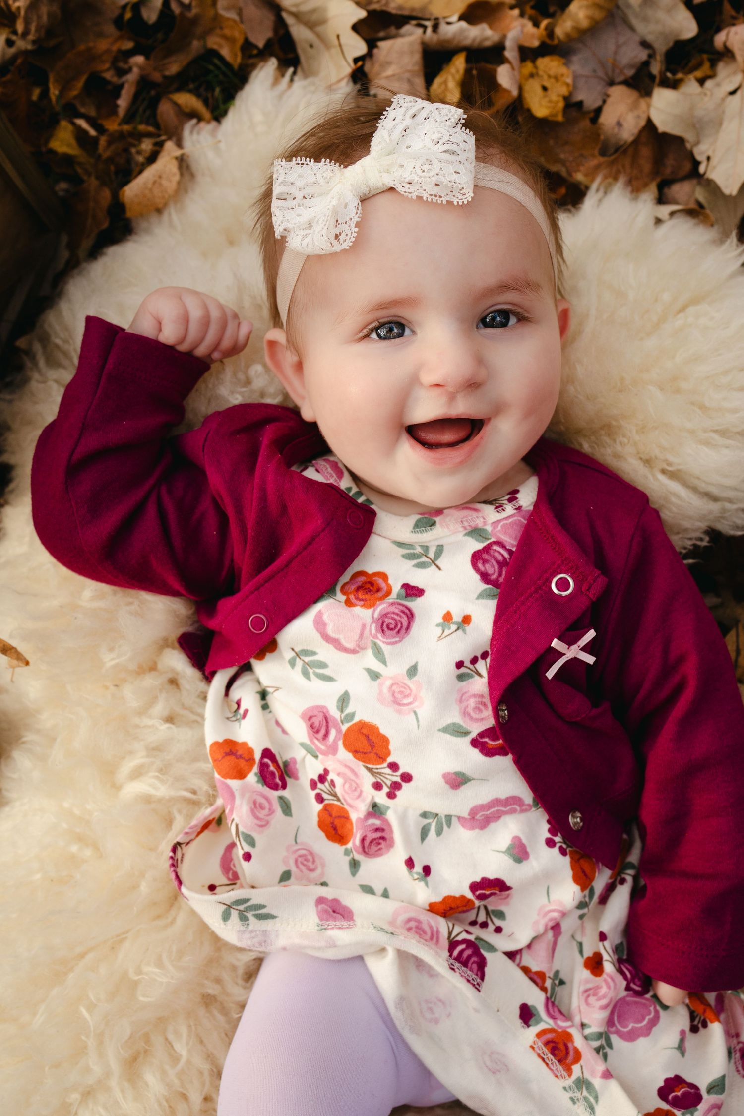 Baby in burgundy cardigan and floral outfit smiling on cream blanket with autumn leaves.