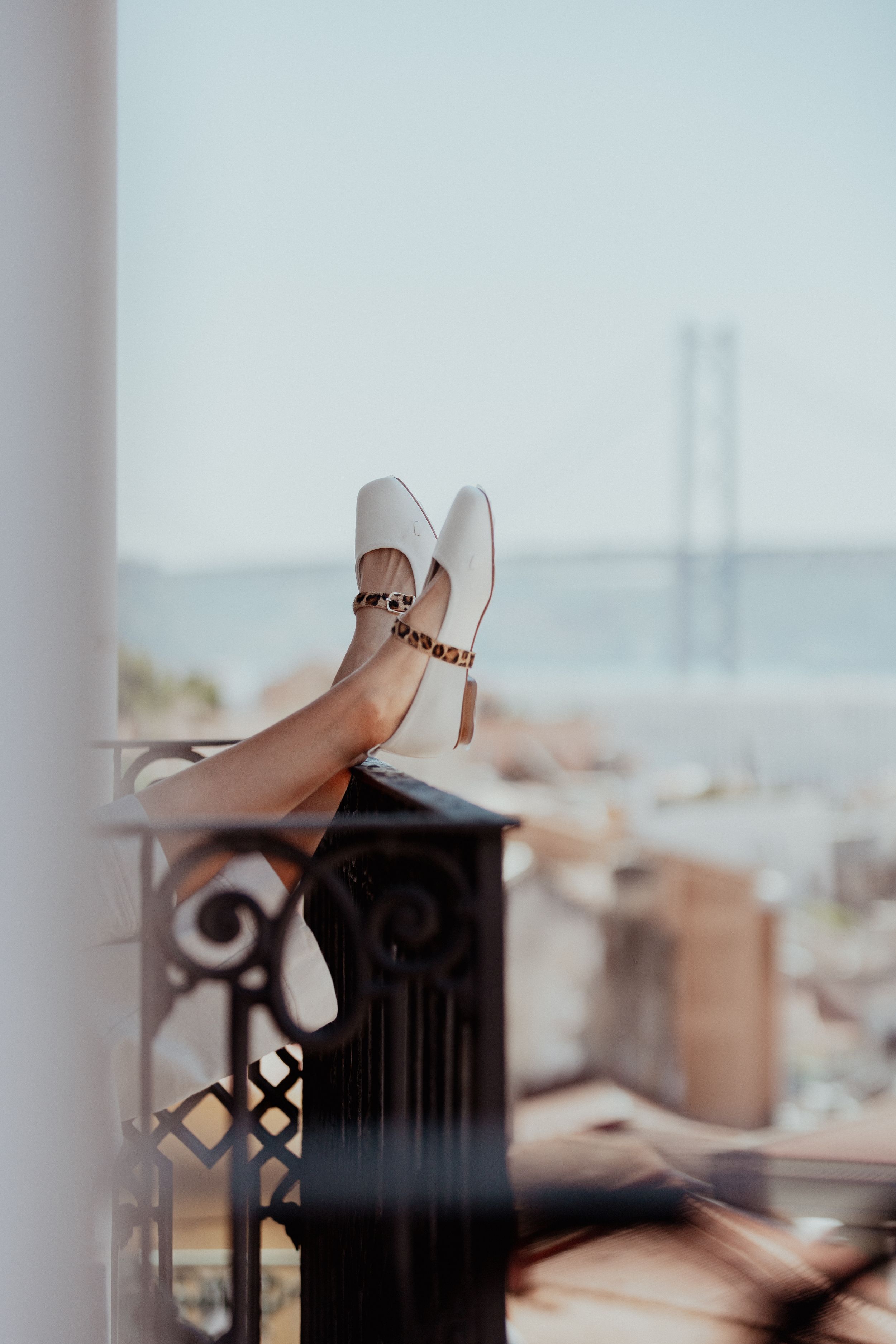 White ballet flats with studded straps propped on balcony railing with bridge view.