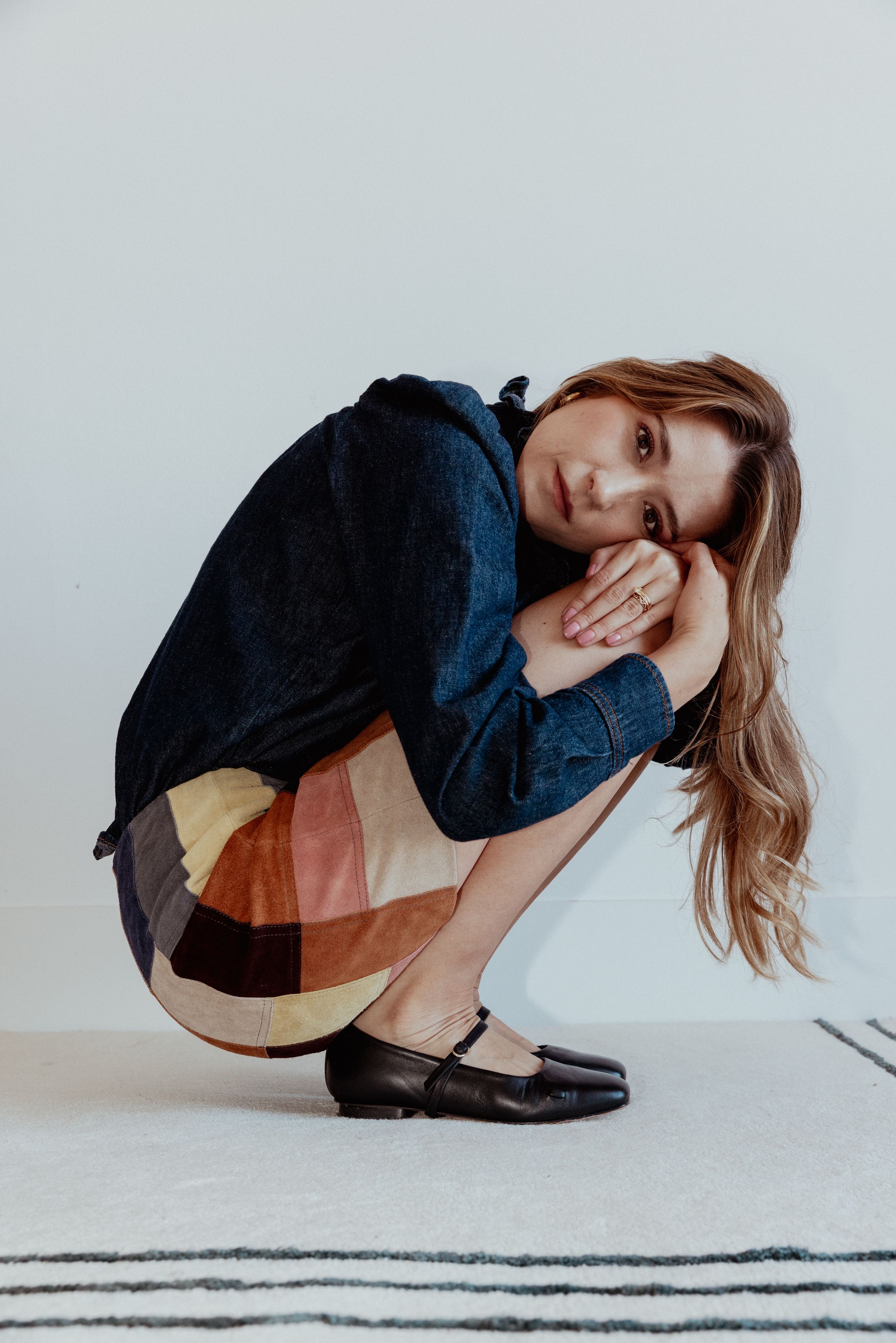 Stylish portrait in navy sweater and patchwork skirt crouching on striped rug.