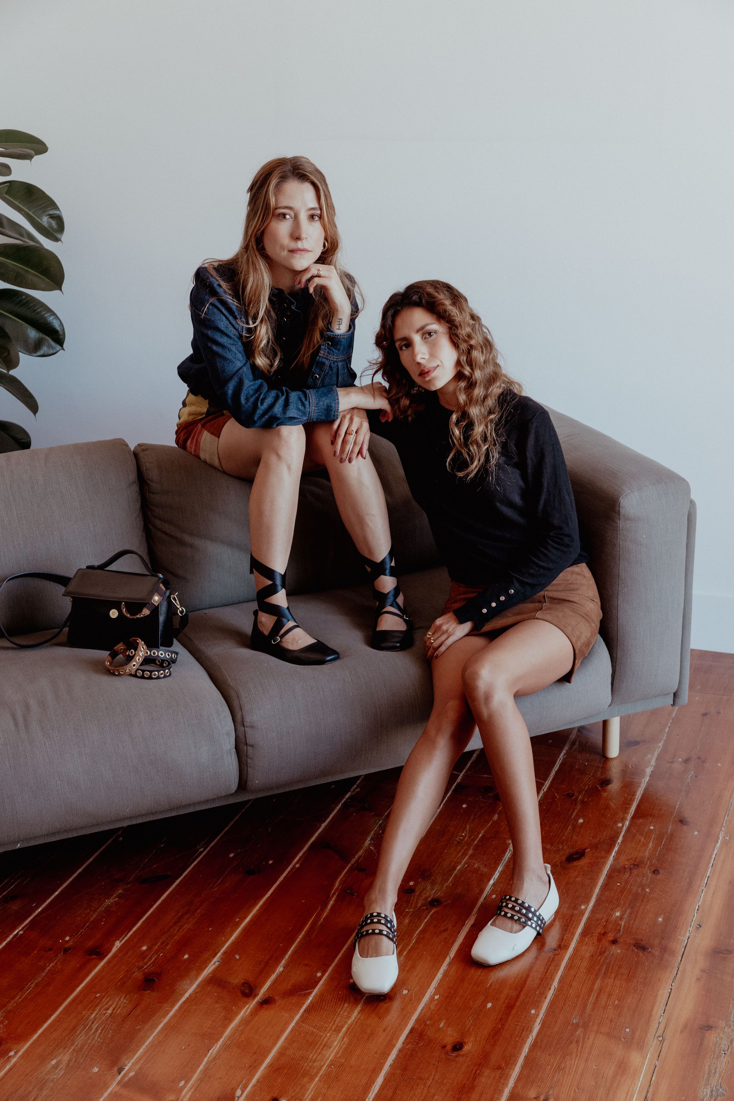 Two women sitting casually on grey sofa wearing dark sweaters and stylish shoes.