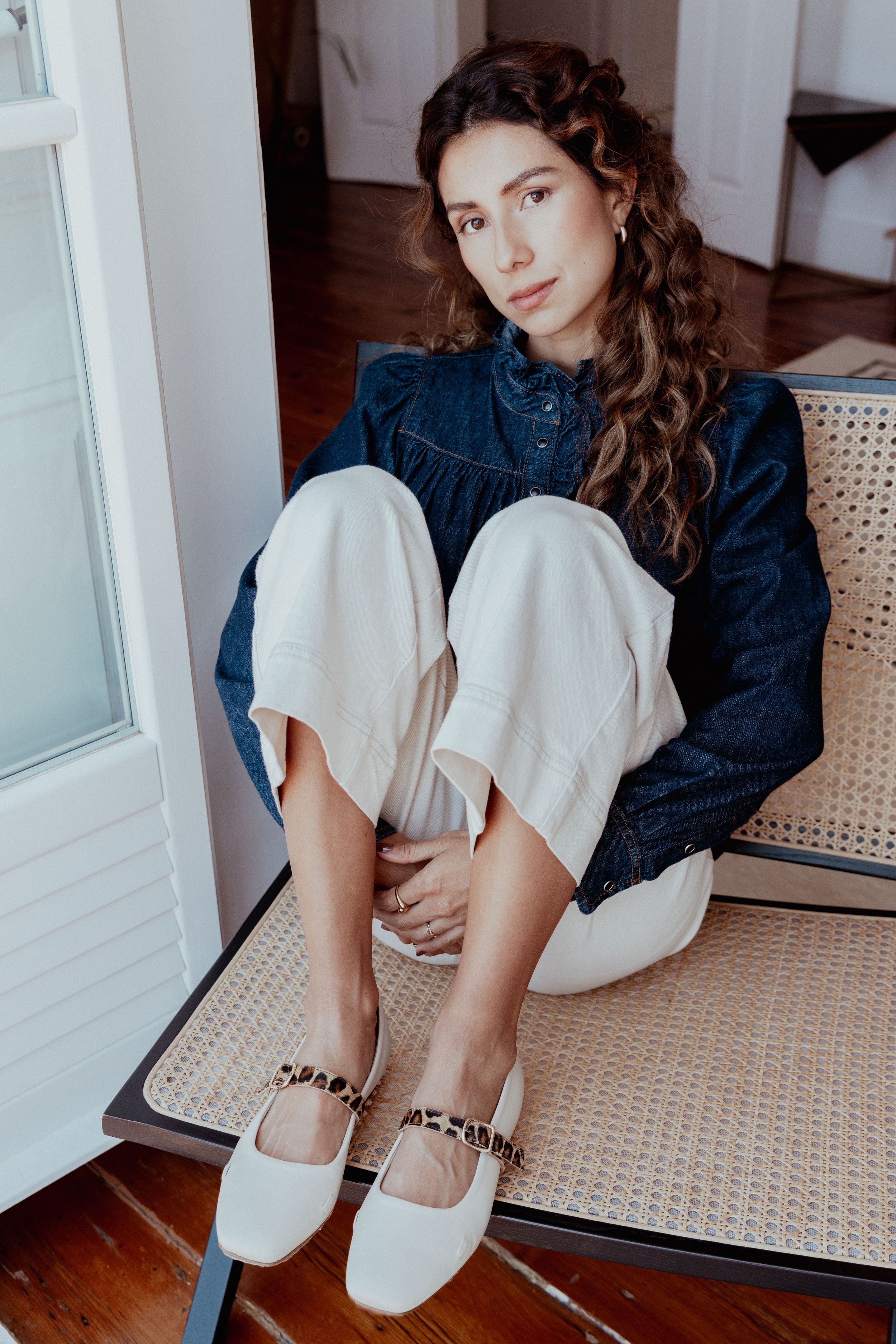 Person in denim shirt and white pants sitting on woven chair showing white ballet flats.