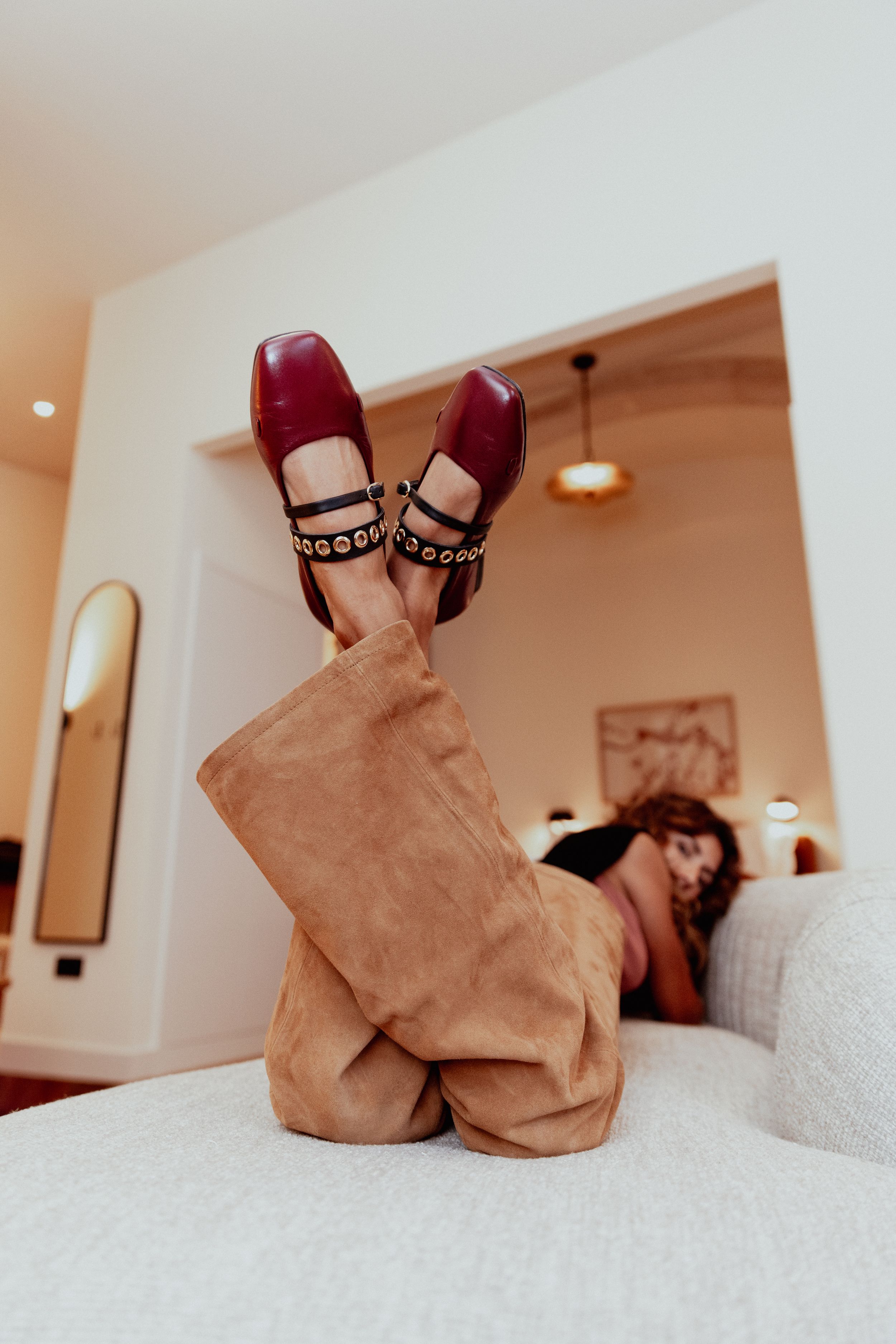 Red mary jane shoes with decorative straps shown on feet resting on a white bed.