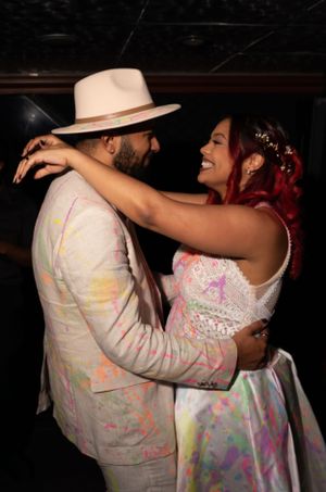 A couple in formal attire splattered with colorful paint dances together at a celebration.