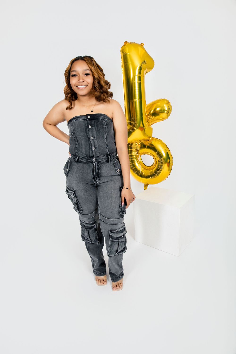 Woman in denim outfit posing with gold number 16 balloons against white background.
