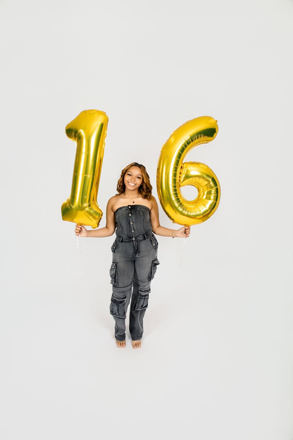 A person poses with large gold number balloons spelling out '16' against a white background.