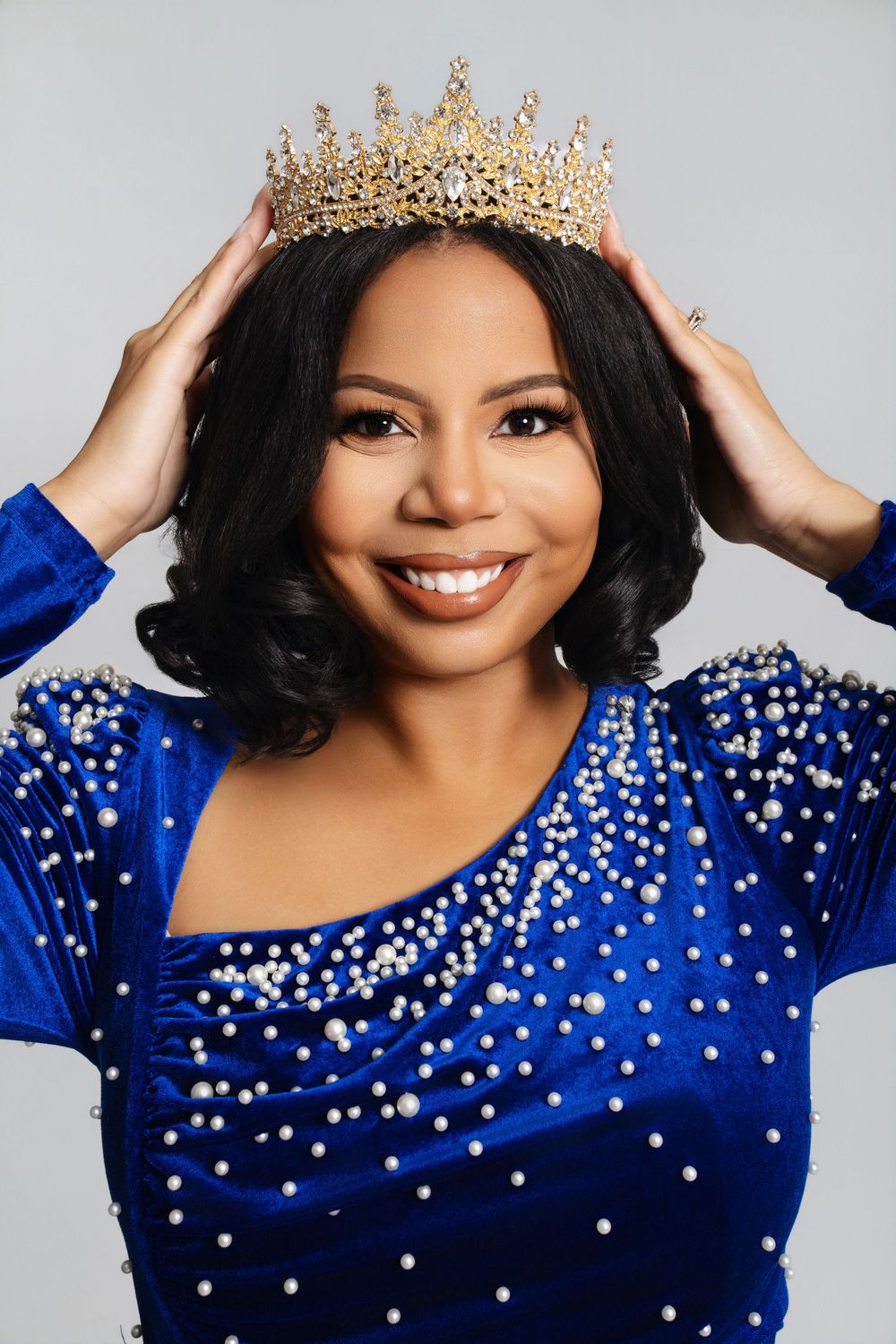 Elegant portrait in royal blue velvet dress adorned with crystals and gold crown against grey background.