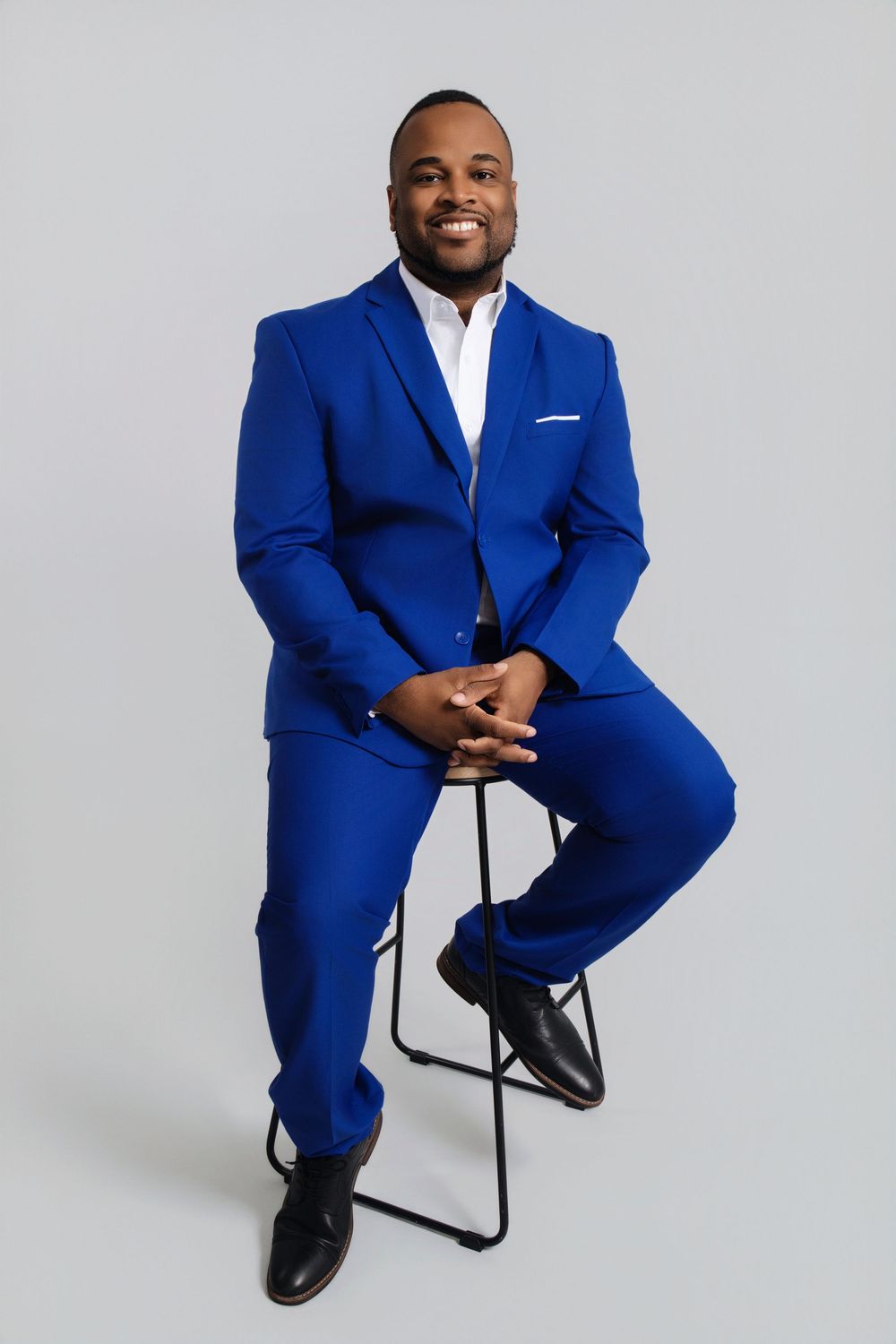 Professional studio portraits of someone wearing a bright royal blue suit while seated on a black stool.