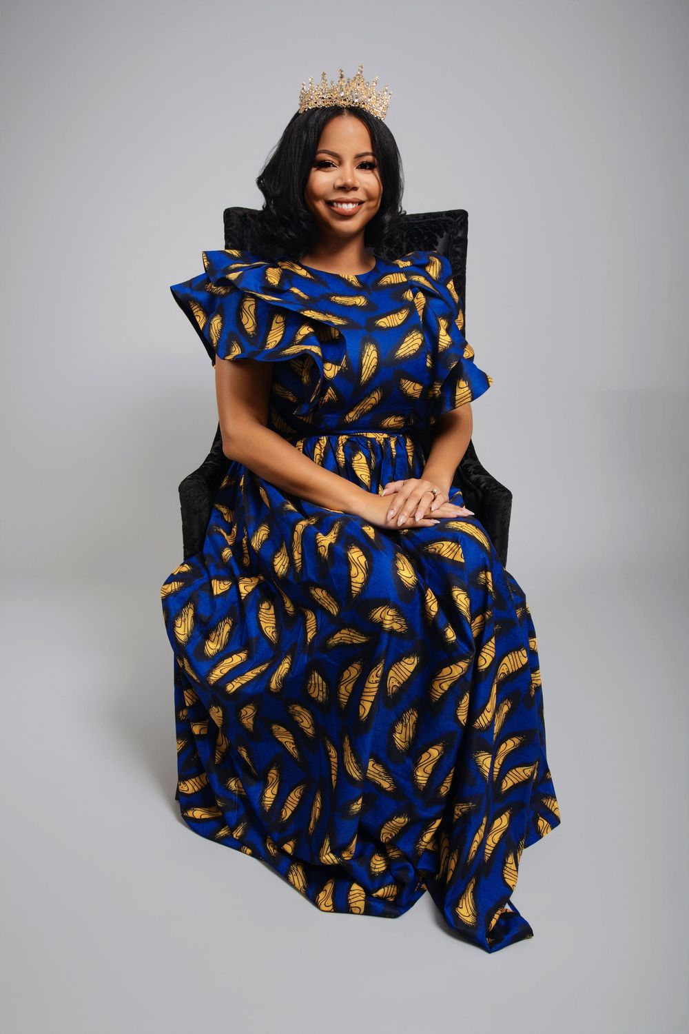 Person in a vibrant blue and gold patterned dress poses elegantly on a chair against a gray backdrop.