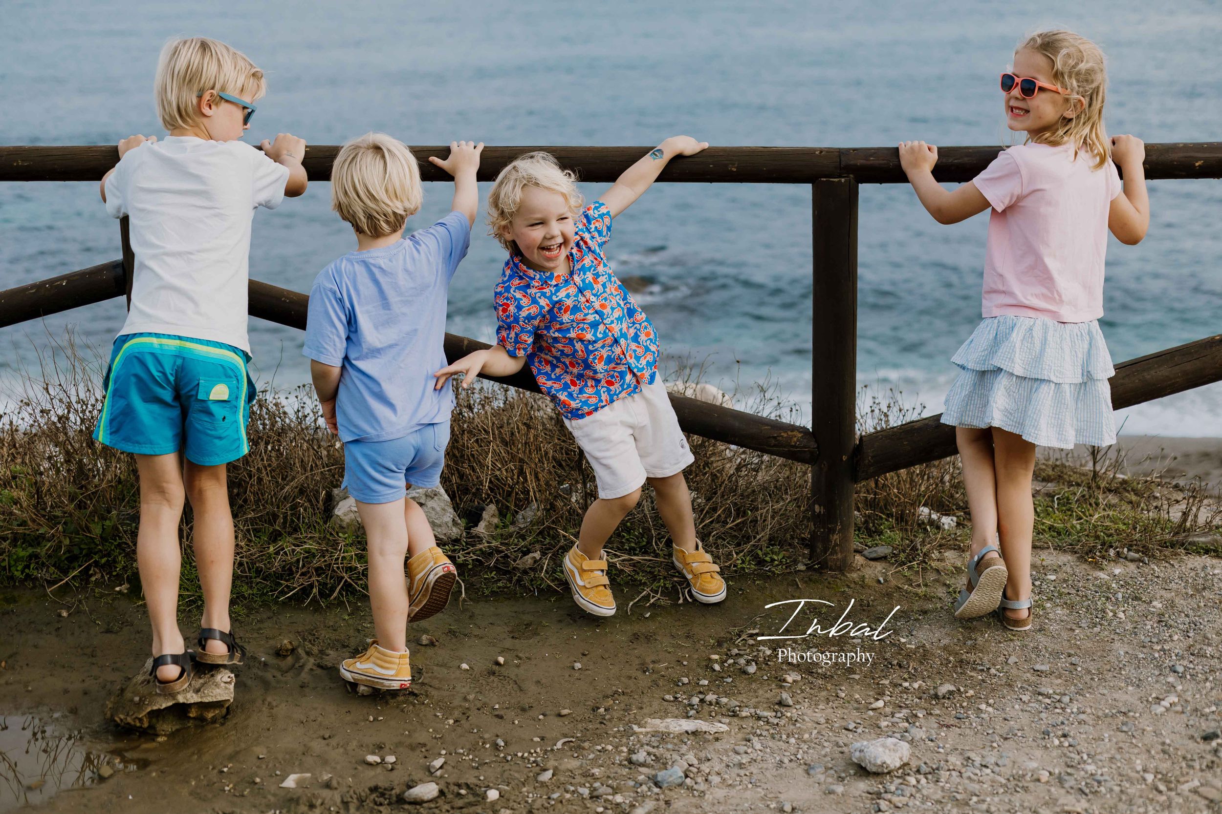 Four young children leaning on a wooden fence overlooking the ocean, with one turning to laugh at the camera.