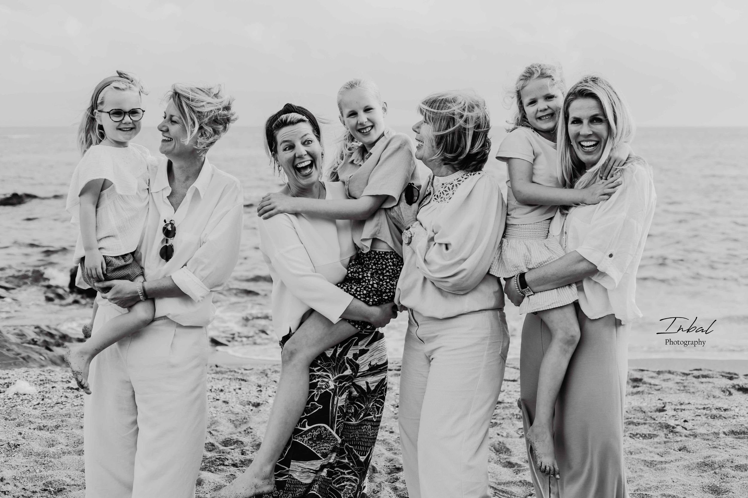 Black and white photo of joyful women and children on a beach. Four women, each holding a child, smile and laugh.