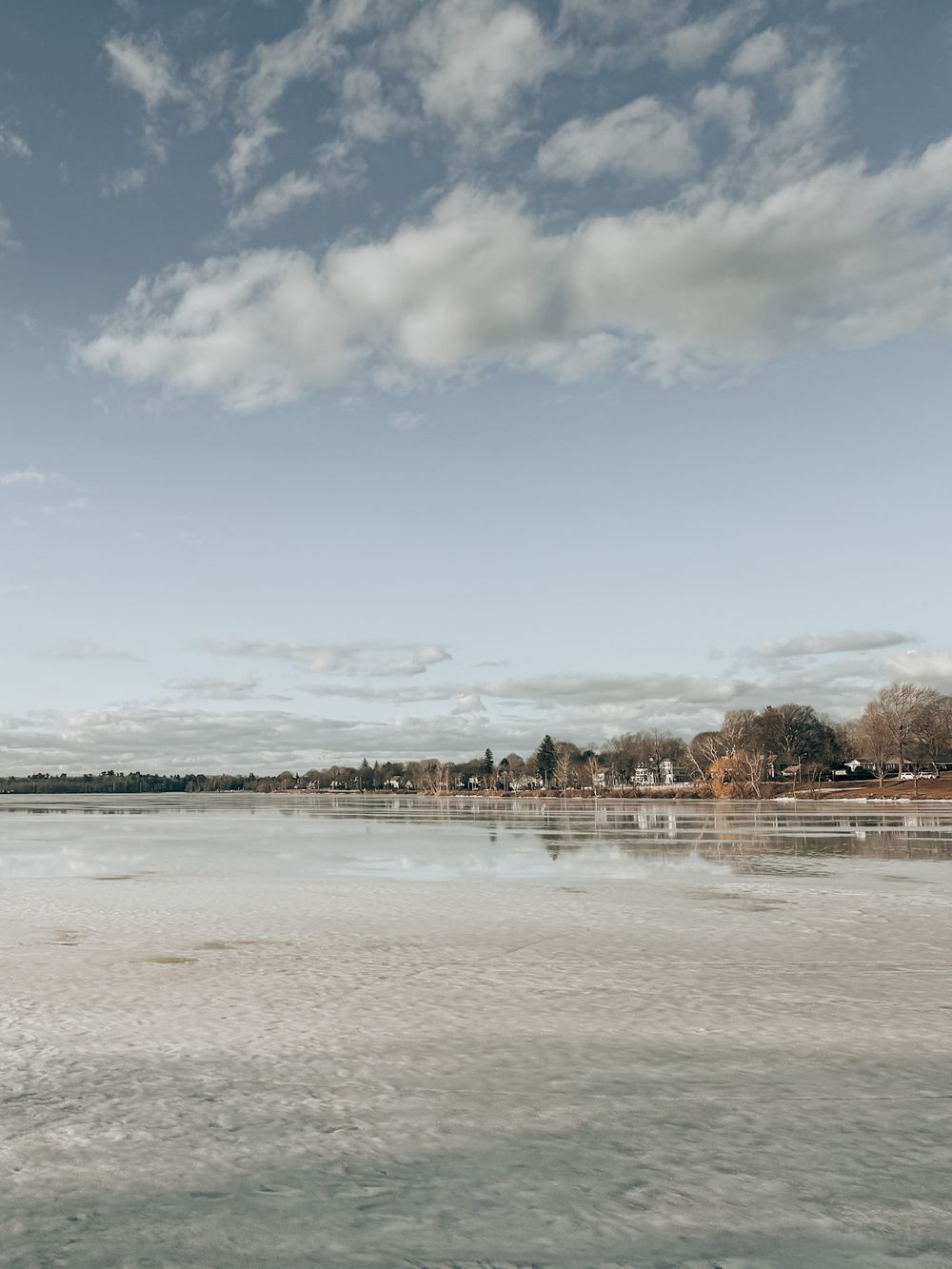 A frozen lake landscape with houses and trees along the shoreline under a cloudy winter sky.