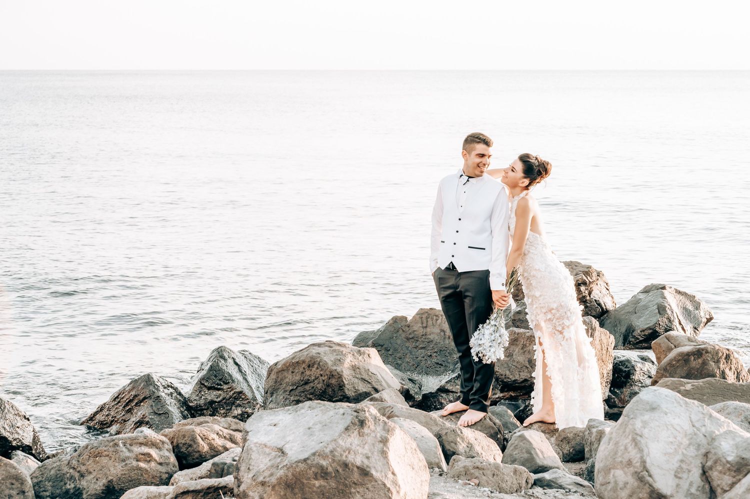 Romantic couple in wedding attire pose on coastal rocks by the sea.