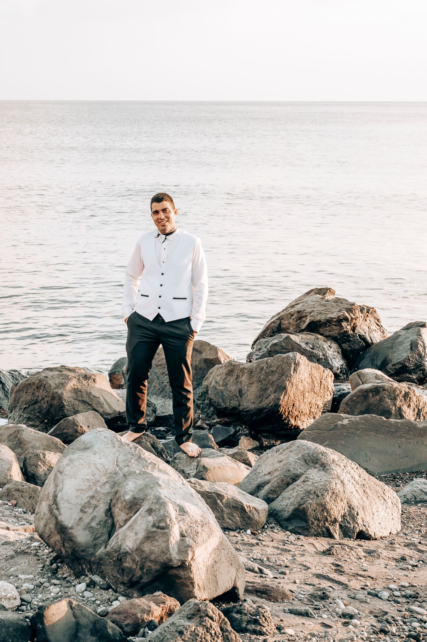 Groom in white vest stands alone on rocky beach coastline.